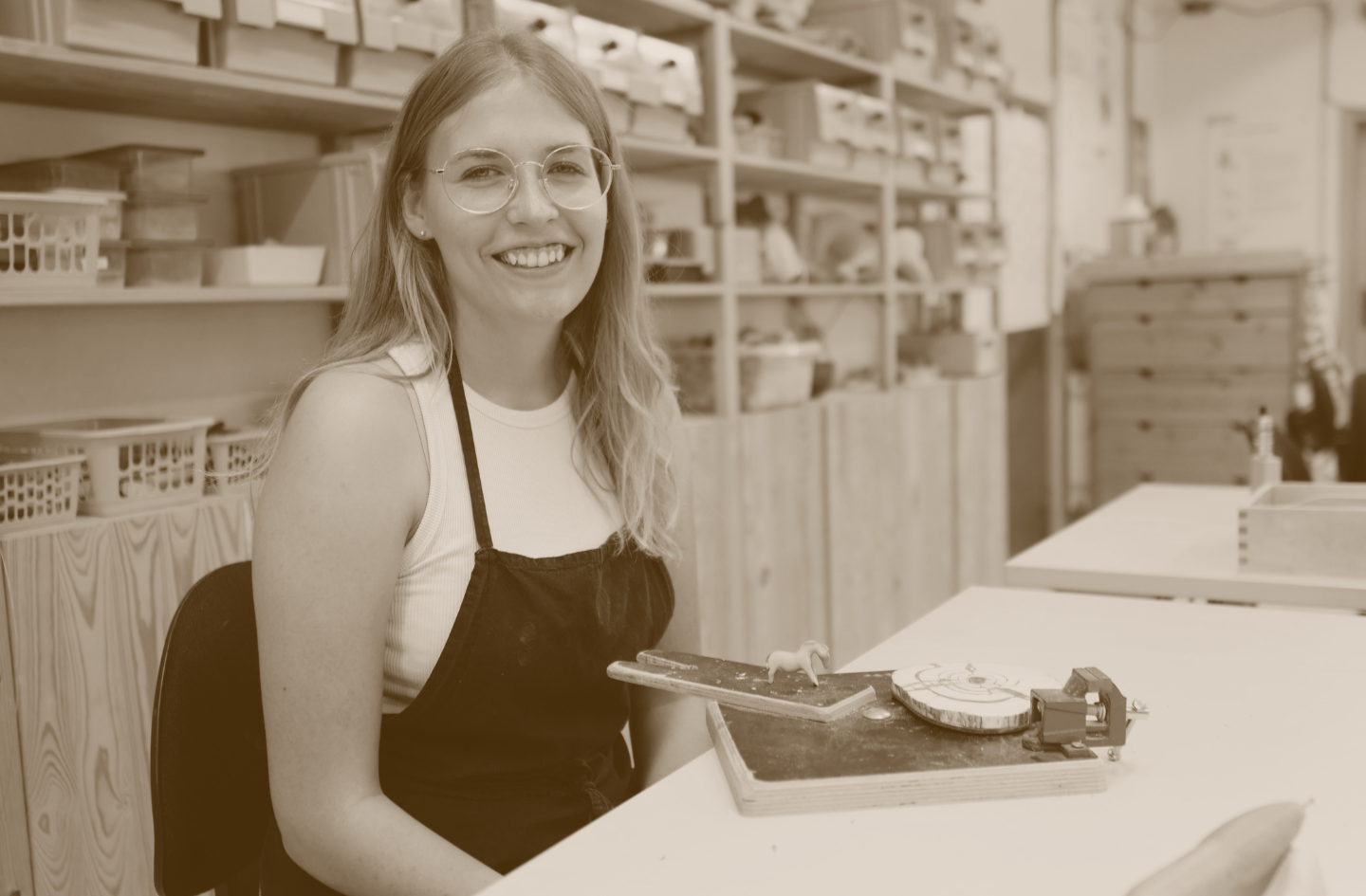 A young woman with long hair and glasses smiling while sitting at a table in an art or craft classroom, with shelves filled with supplies in the background.