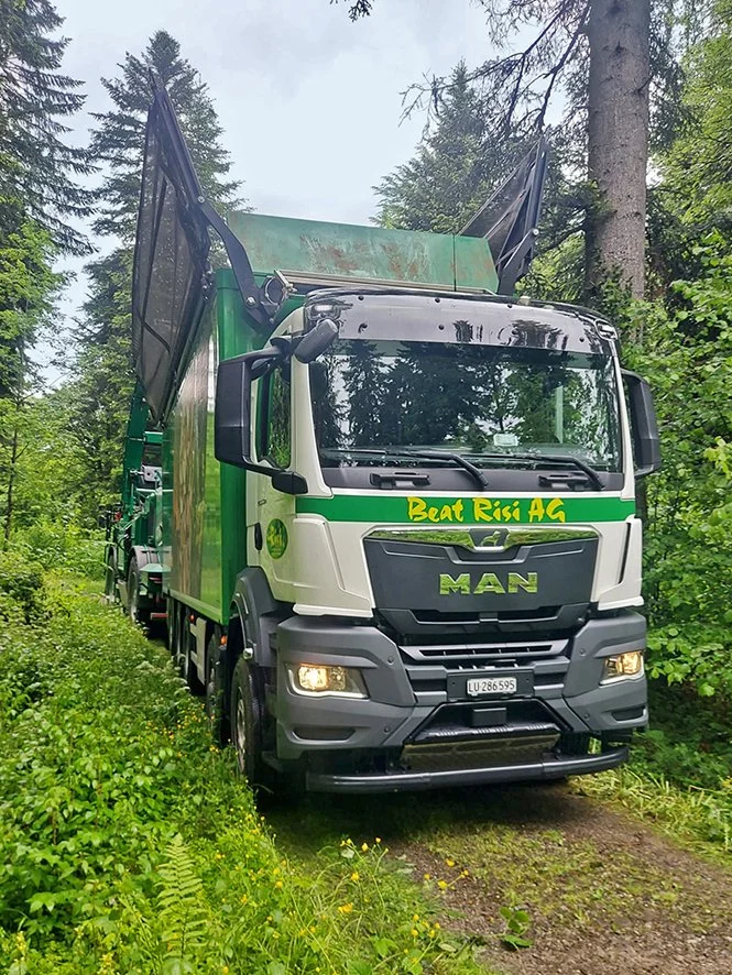 Ein großer MAN-LKW steht auf einem schmalen Weg in einem grünen Wald, mit geöffneten Müllcontainern auf dem Lkw.