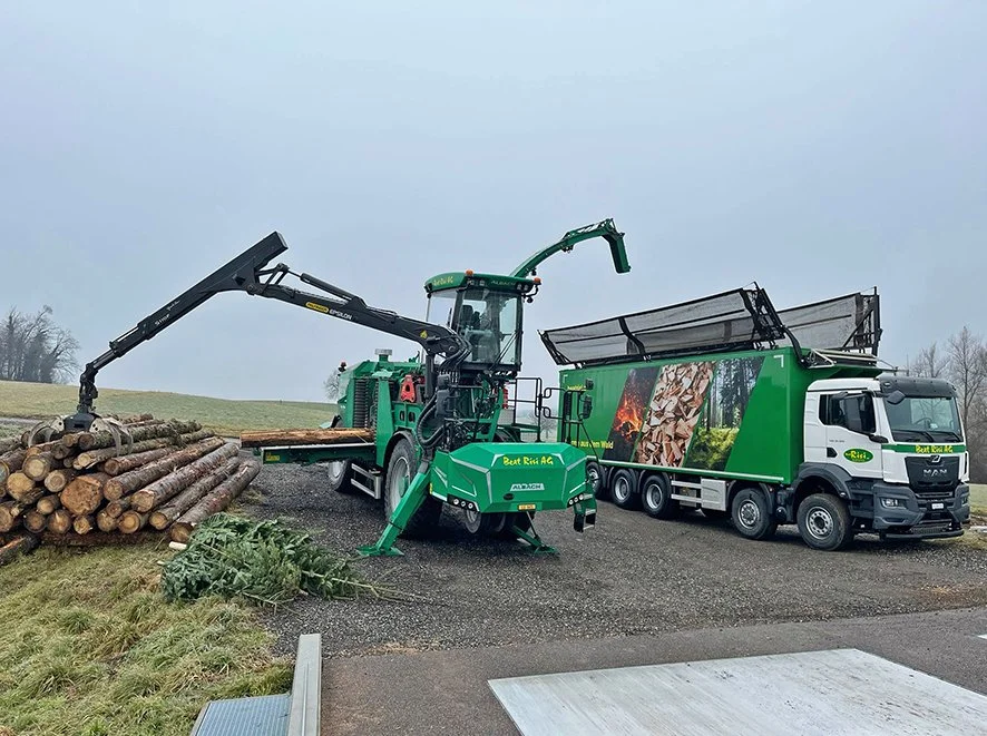 Holzerntemaschine beim Aufnehmen von Baumstämmen, daneben ein umgebauter LKW mit Werbung für eine Firma im Holz- und Forstsektor, auf einem Feld bei bedecktem Himmel.