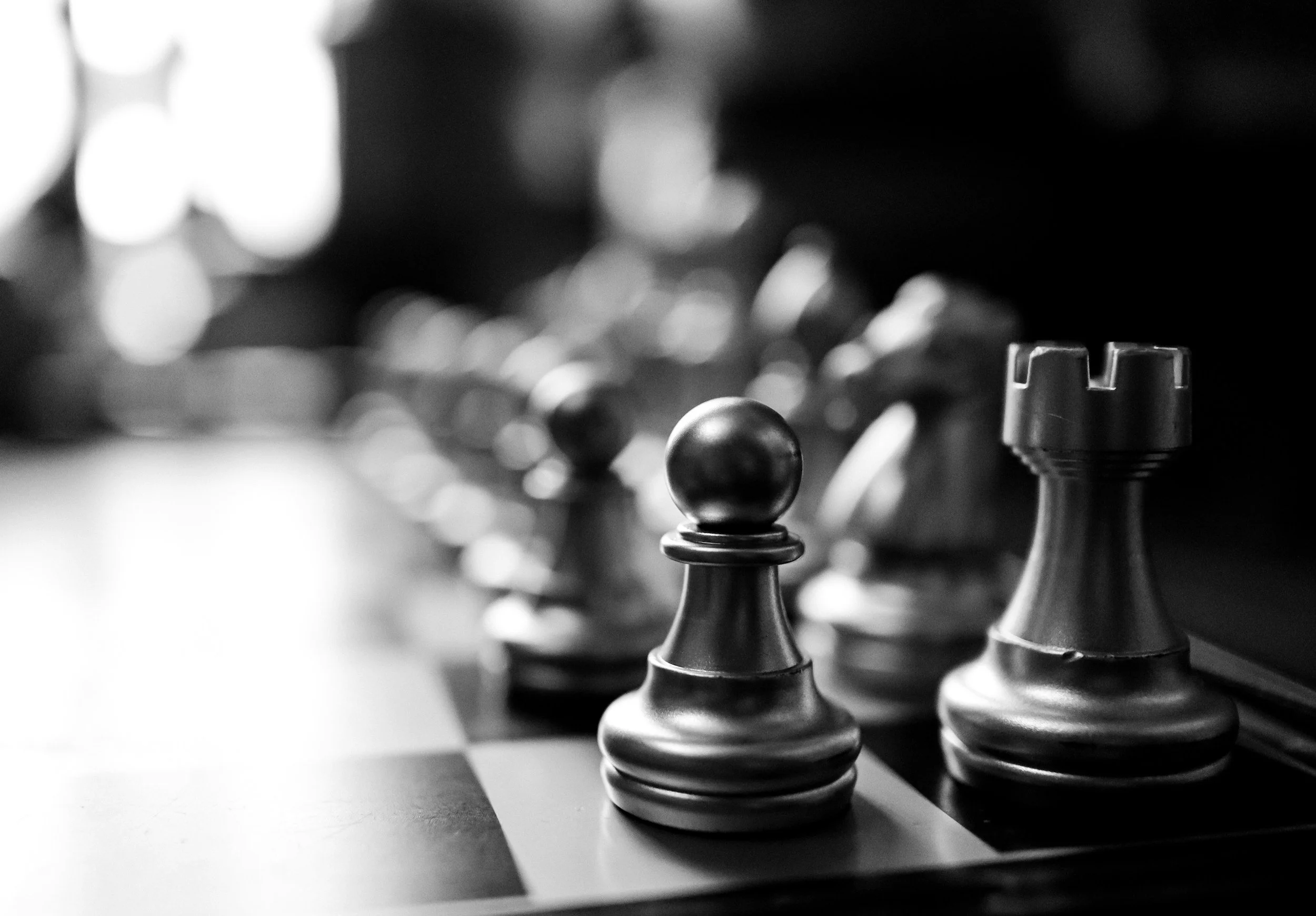 Schwarze und weiße Schachfiguren auf einem Schachbrett, im Vordergrund ein Springer und ein Bauer im Fokus, im Hintergrund sind unscharfe, beleuchtete Figuren.