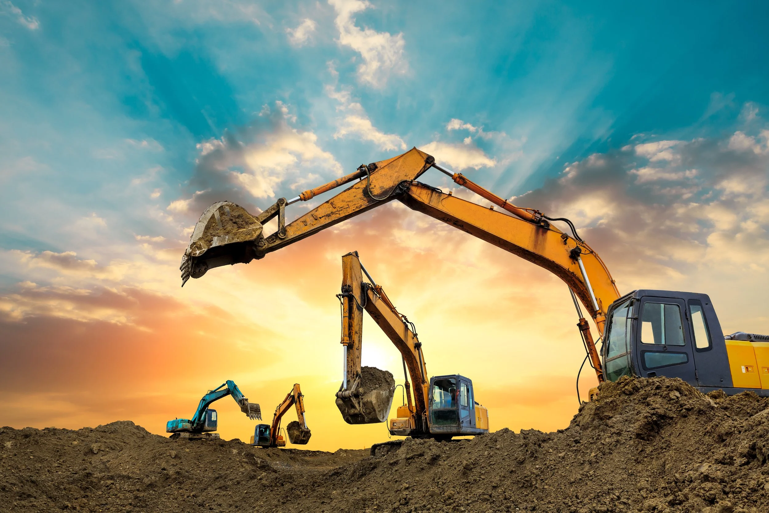 Several construction excavators working on a dirt site with a sunset sky in the background