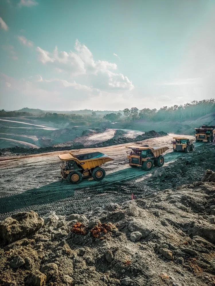 Heavy construction trucks moving dirt on a large, open construction site under a partly cloudy sky.