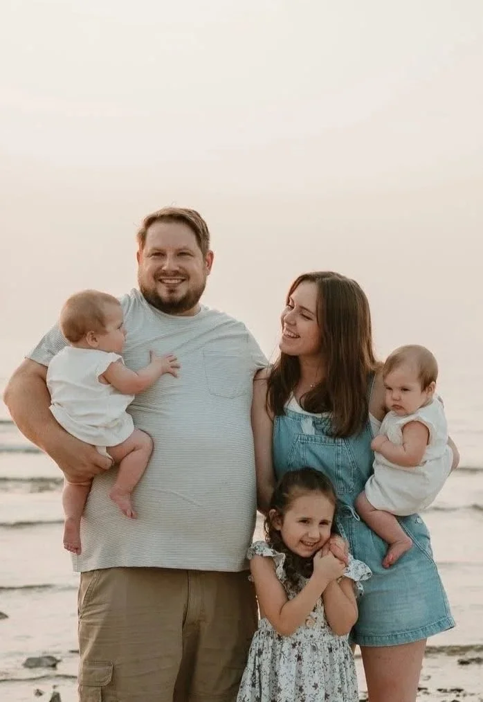 A family of five on the beach during sunset, smiling and looking at each other. The father is holding a baby girl on his left arm, and the mother is holding a toddler girl in her arms. A young girl stands in front, smiling and hugging the mother.