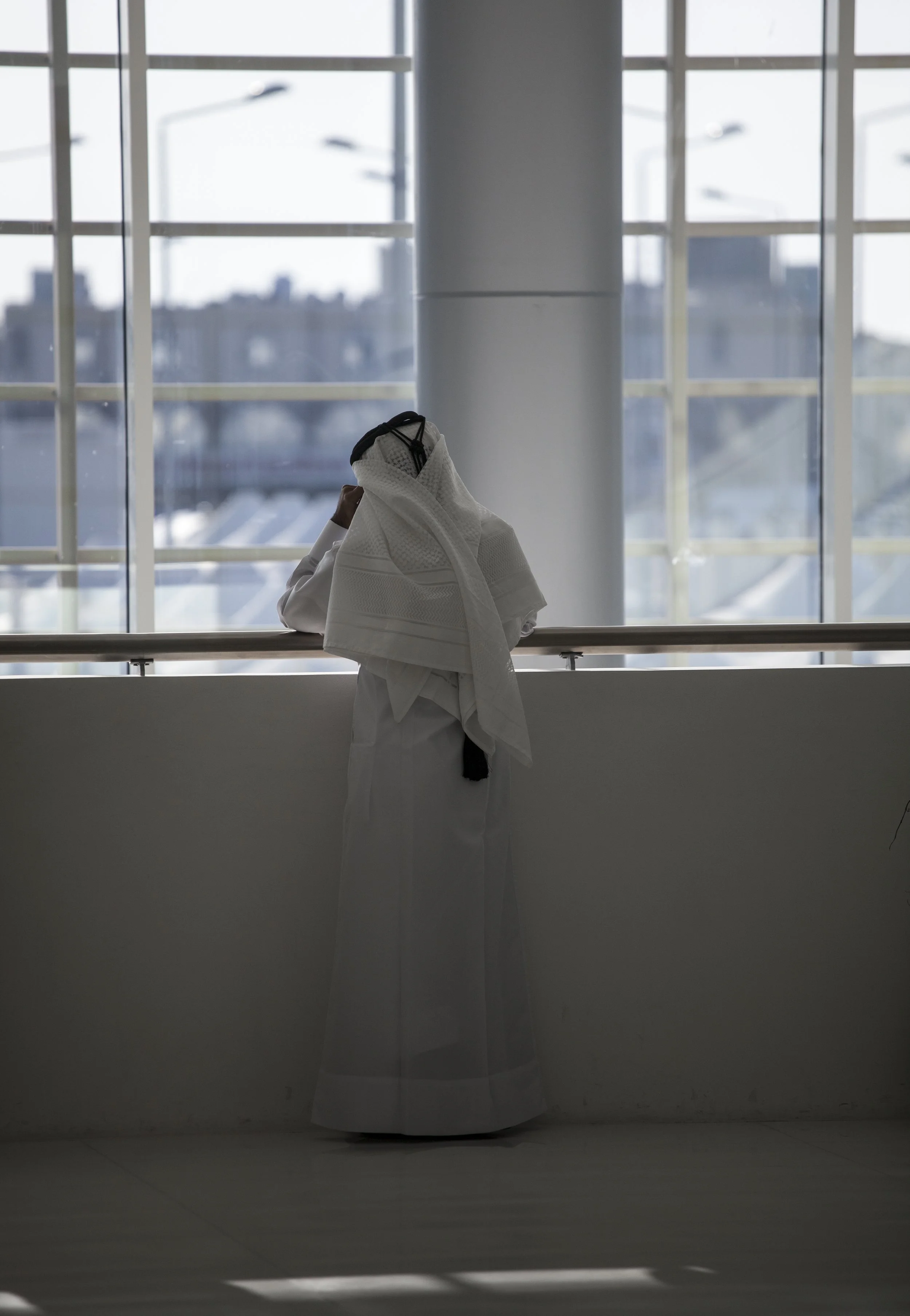 Une personne en kimono blanc se tient près d'une grande fenêtre avec une vue urbaine en arrière-plan, sa tête est recouverte d'un vêtement blanc.