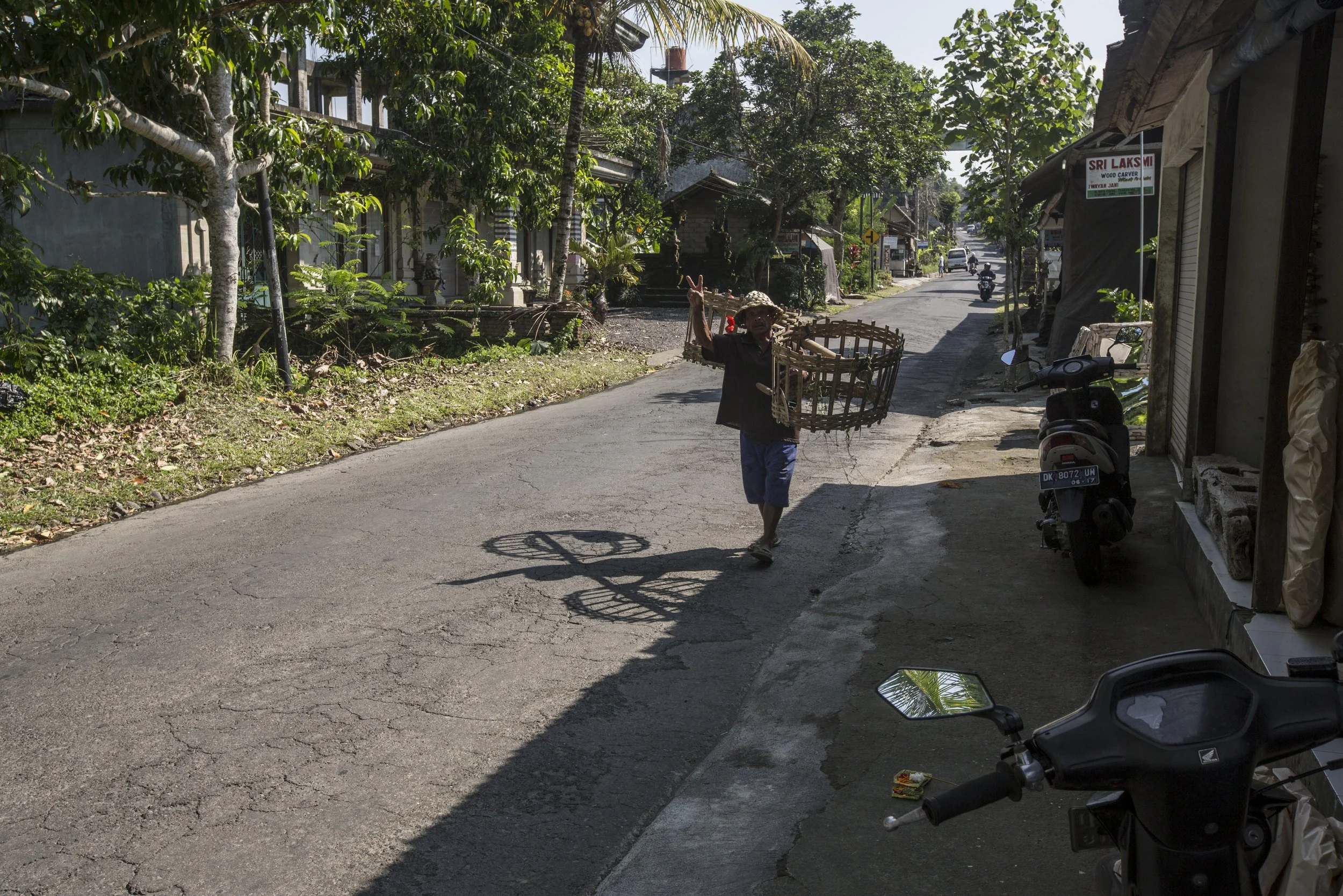 Une femme avec un chapeau porté de côté marche dans une rue pavée tout en tenant une grande cage en bambou, faisant un signe de paix avec la main. La rue est bordée de maisons avec beaucoup de végétation, et il y a une moto stationnée sur le côté, av