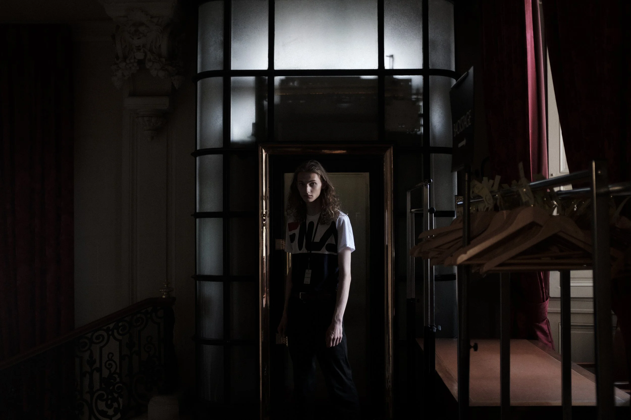 Jeune femme debout dans une pièce sombre, regardant vers l'objectif, derrière une verrière, avec un éclairage faible et des vêtements suspendus à une grille.