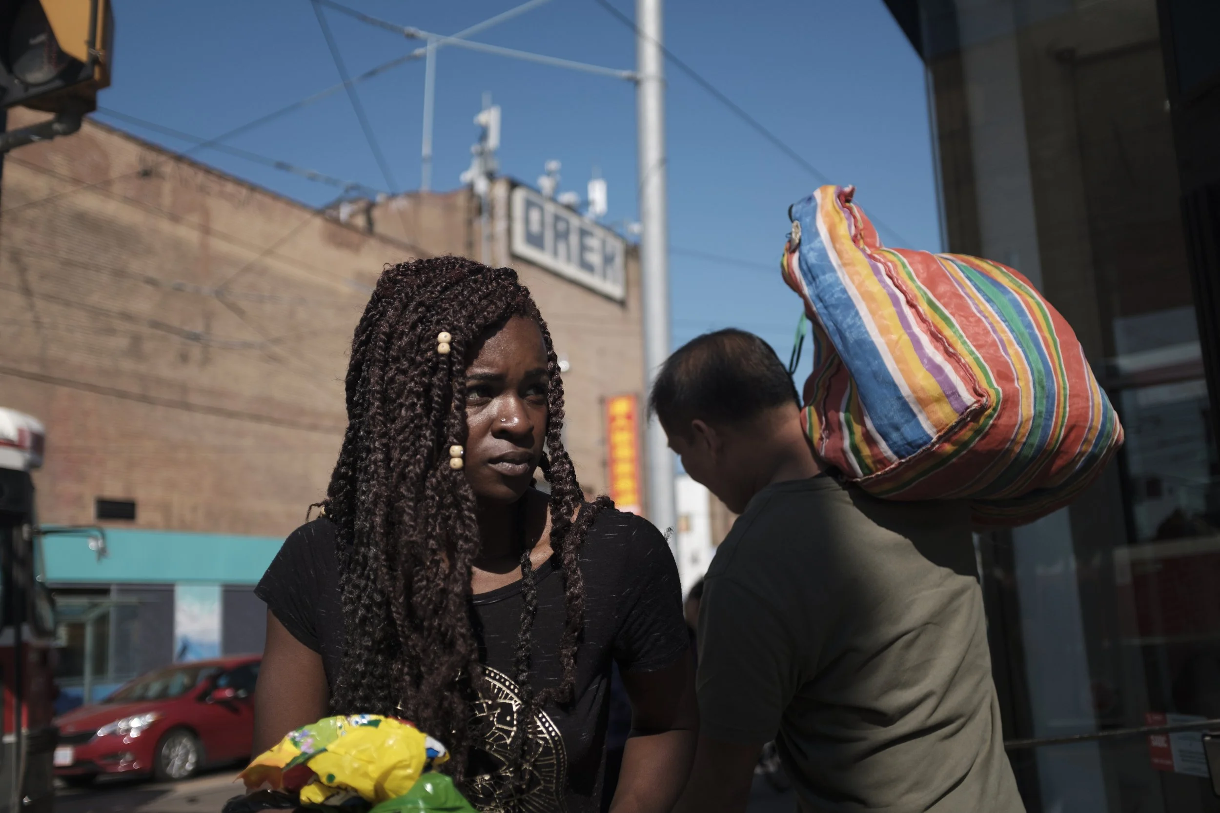 Une femme aux cheveux bouclés et une homme portant un sac coloré sur l'épaule se trouvent sur une rue en pleine journée, avec un bâtiment en arrière-plan. 