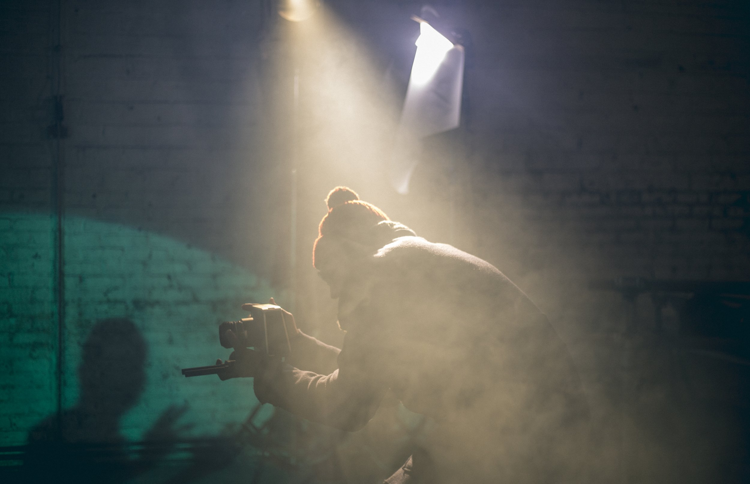 Une personne portant un bonnet rouge et une veste sombre, tenant une caméra dans une pièce sombre avec une lumière venant d'une fenêtre au plafond.