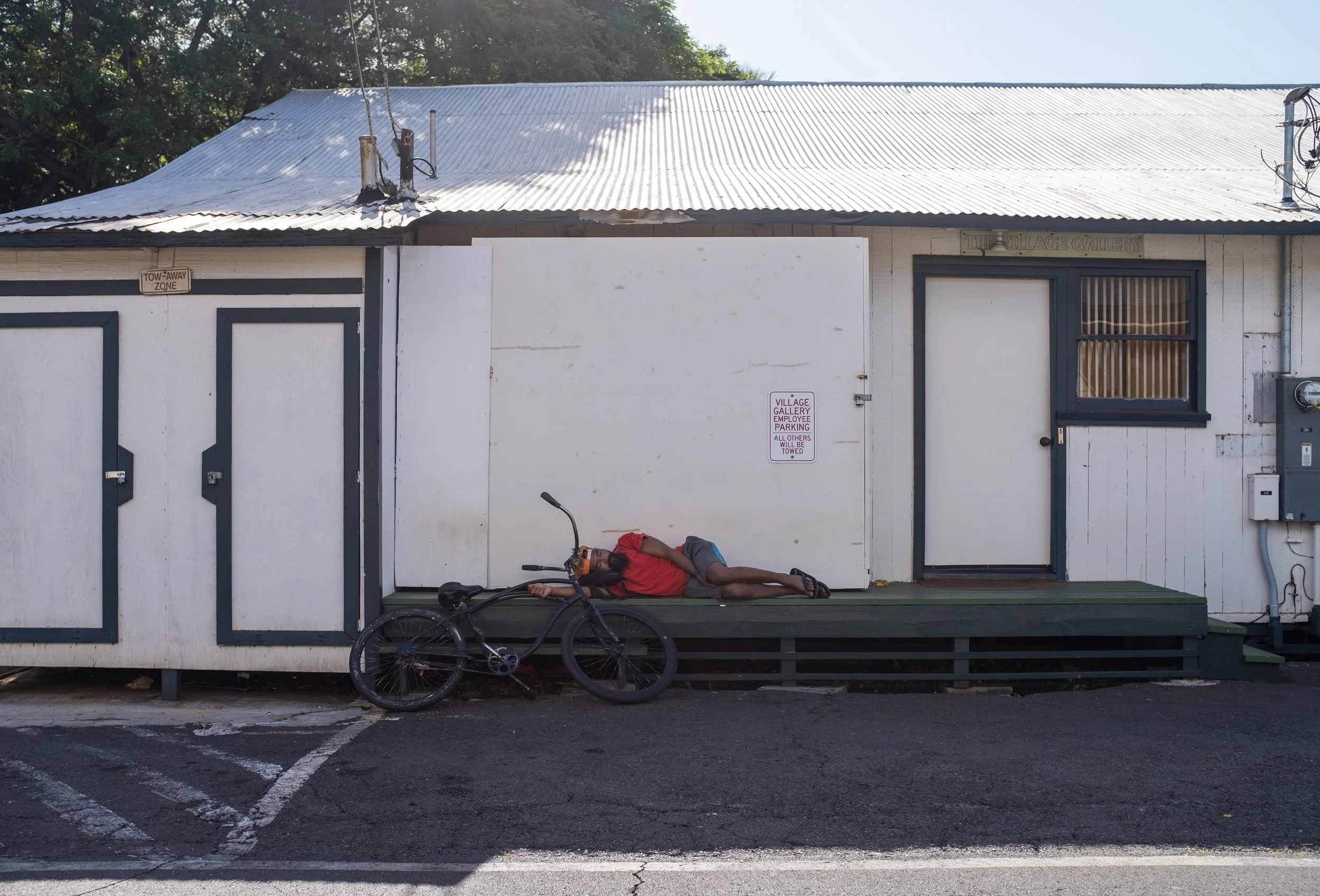 Une personne allongée sur un trottoir devant un bâtiment en bois blanc, avec un vélo garé à côté, sous un ciel clair.