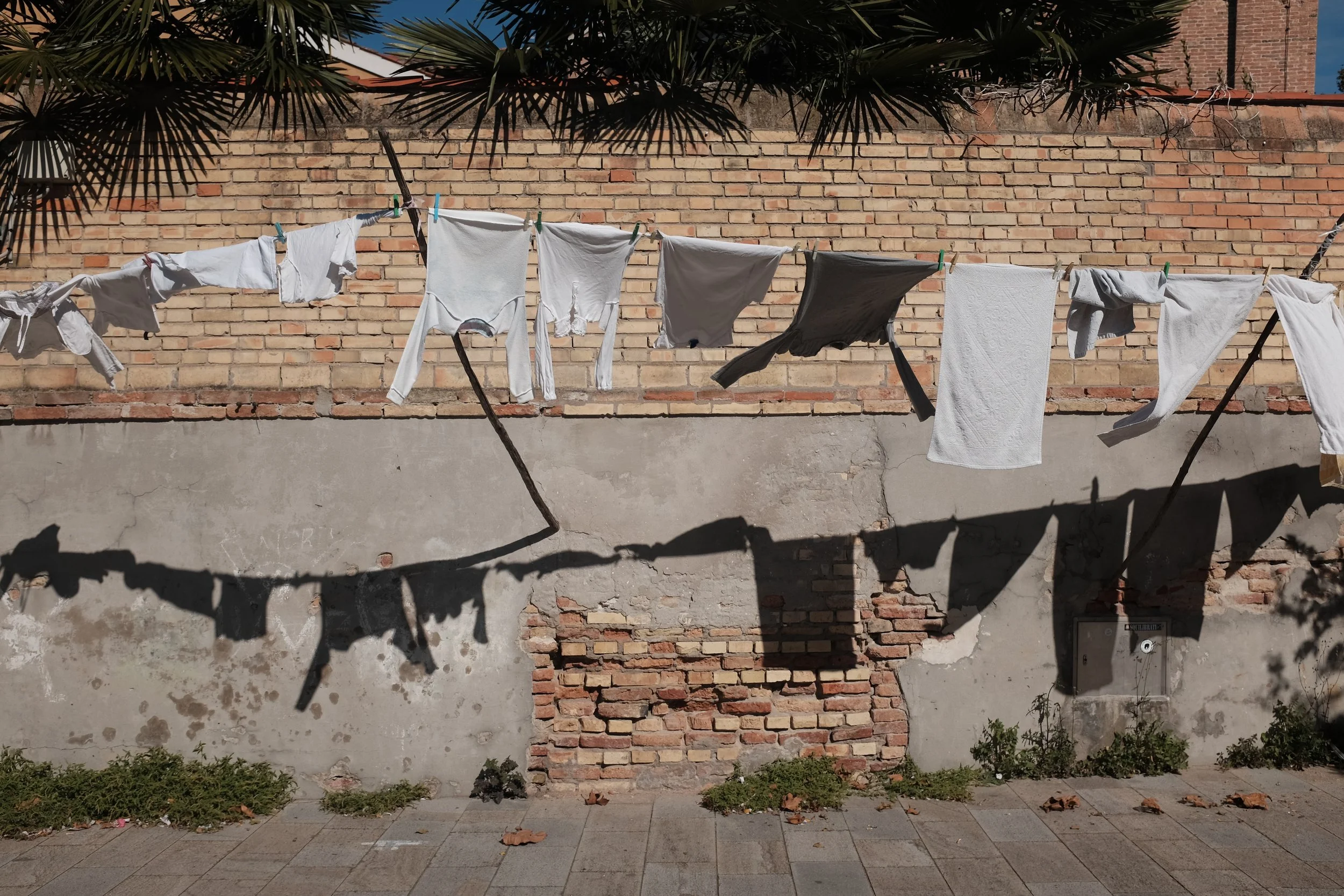 Linge blanc séchant sur une corde à linge, avec ombres projetées sur un mur en briques et en béton, sous un palmier.