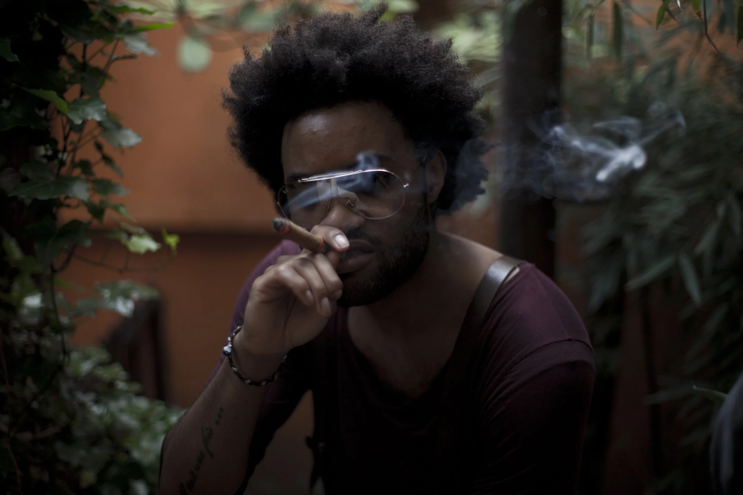 Un homme avec des cheveux bouclés portant des lunettes de soleil fume un cigare dans un environnement extérieur entouré de végétation.