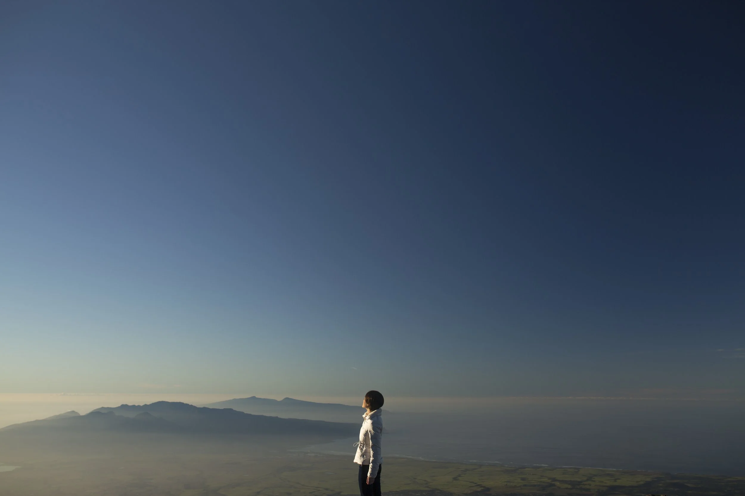 Une femme se tenant sur une colline avec une vue panoramique, regardant au loin vers des montagnes et la mer au coucher du soleil, sous un ciel clair.