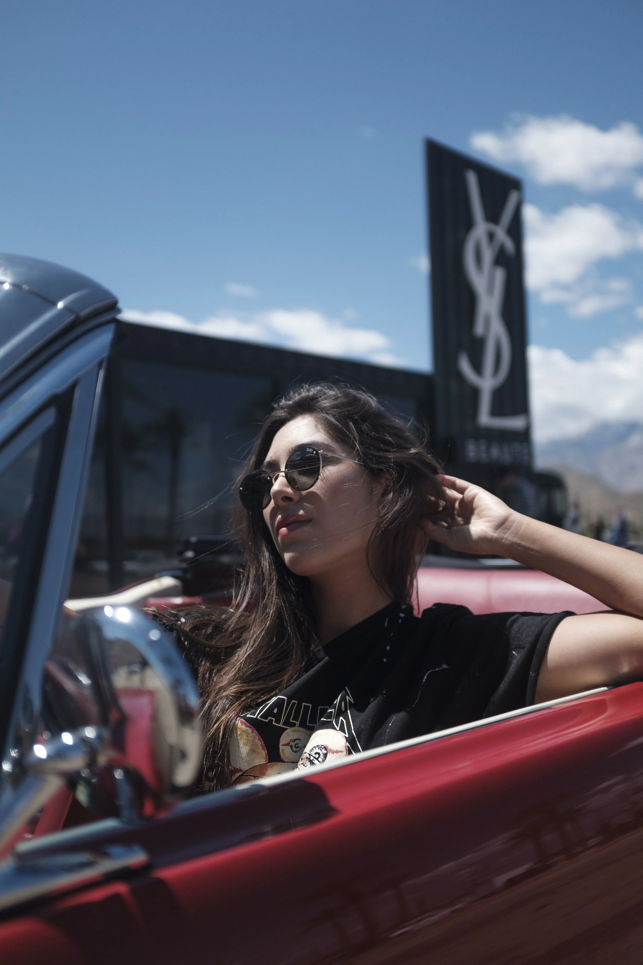 Jeune femme avec des lunettes de soleil, assise dans une voiture décapotable rouge, sous un ciel bleu avec quelques nuages, avec un panneau publicitaire en arrière-plan.