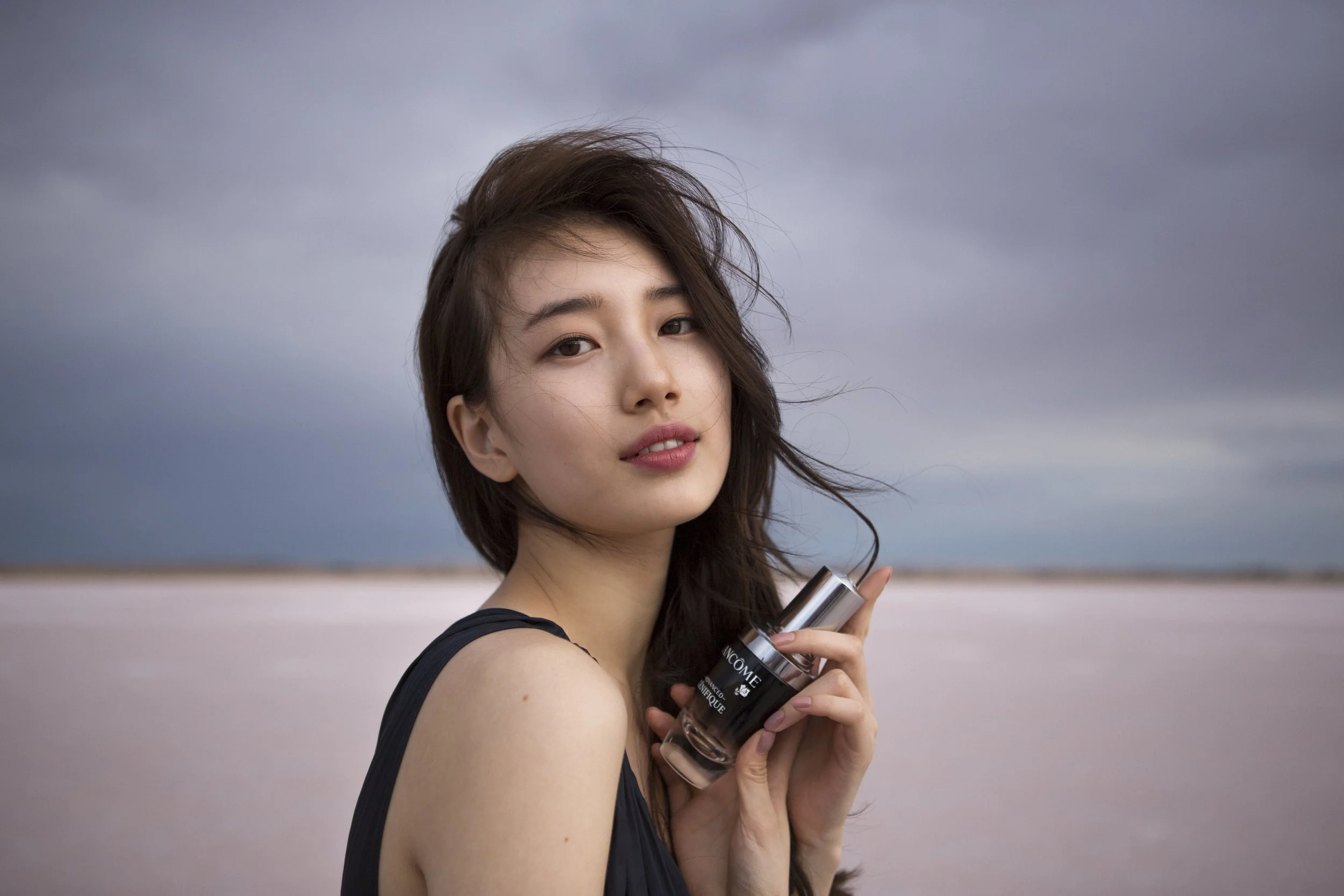 Une femme avec des cheveux bruns, tenant un flacon de produit cosmétique de la marque Lancôme, devant un paysage naturel avec un ciel nuageux.