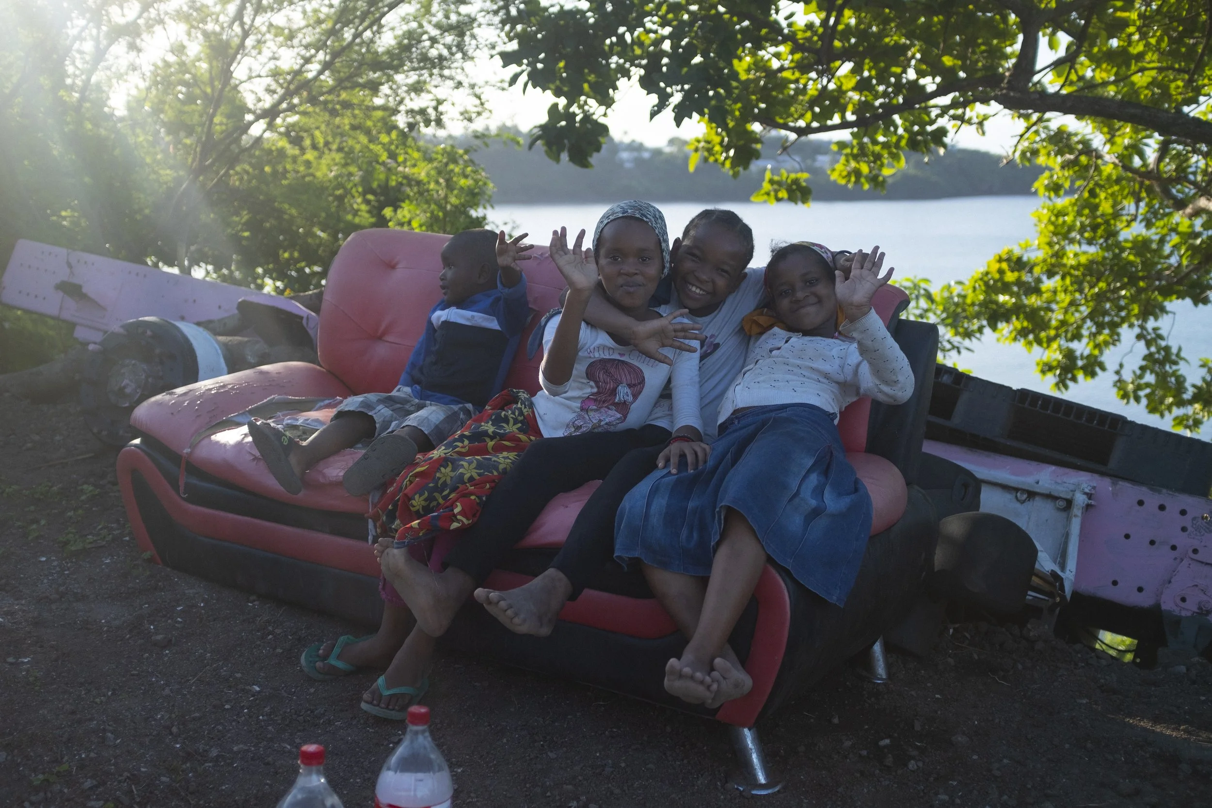 Quatre enfants souriants assis sur un ancien canapé rouge en plein air, entourés d'arbres et à côté d'un lac. Deux enfants font signe de la main, tous semblent heureux.