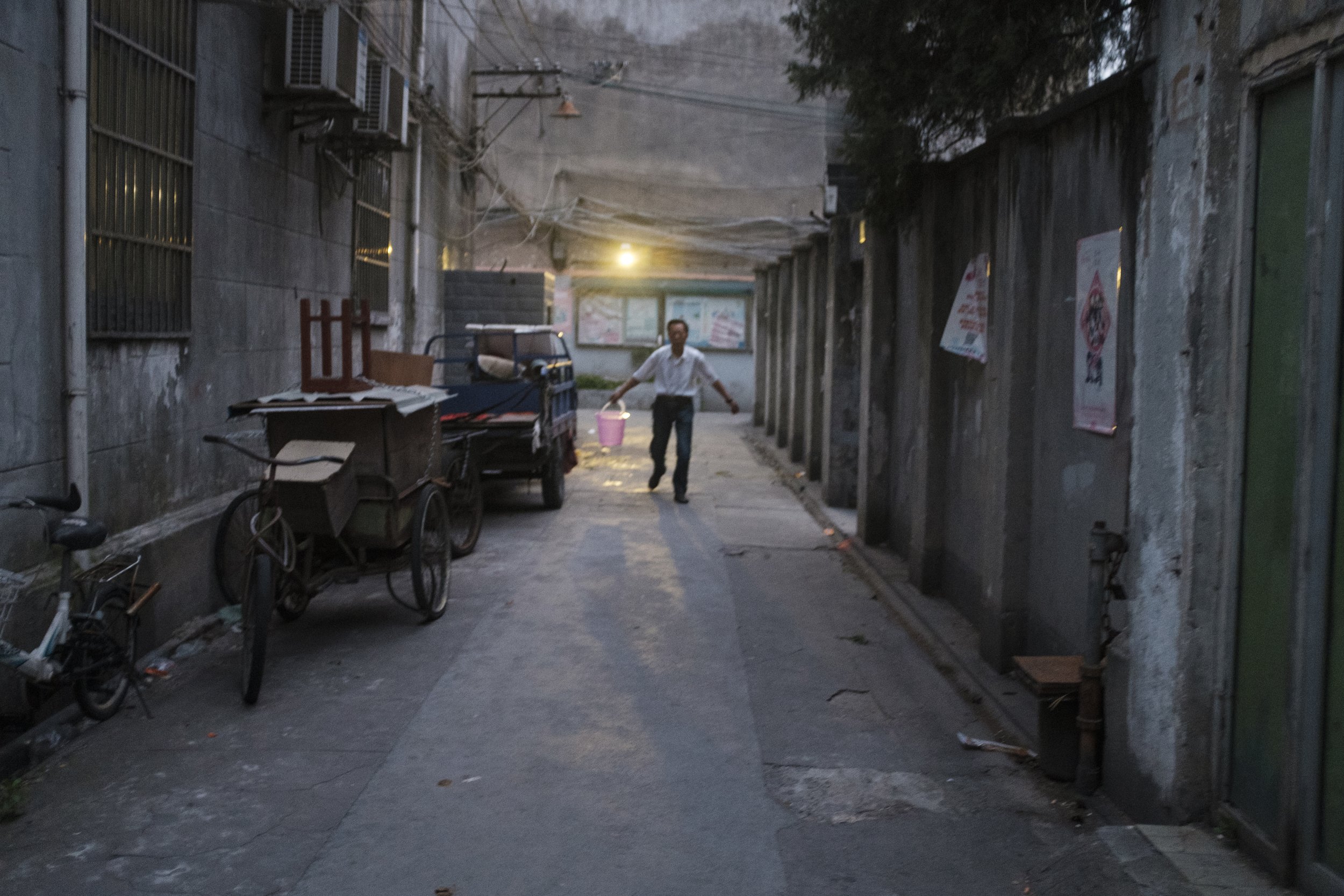 Une ruelle urbaine en fin d'après-midi avec un homme marchant, tenant un seau rose, entouré de vieux bâtiments, vélos et crates de livraison.