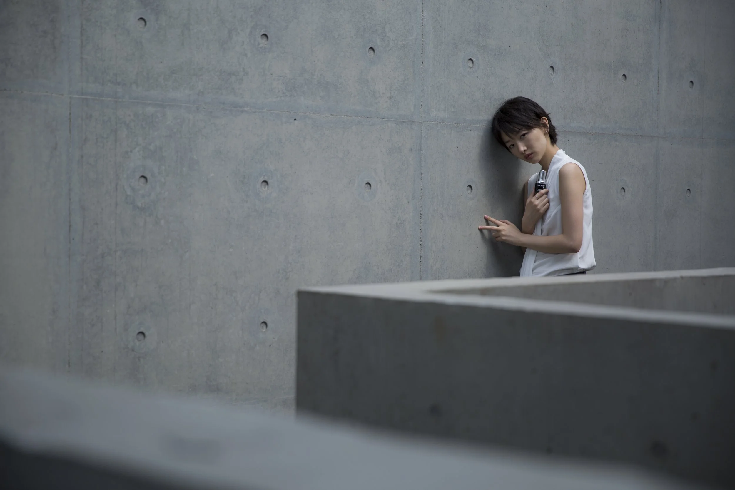 Une jeune femme porte une chemise blanche sans manches, se tient contre un mur en béton gris, fait un signe de la paix et regarde l'objectif.