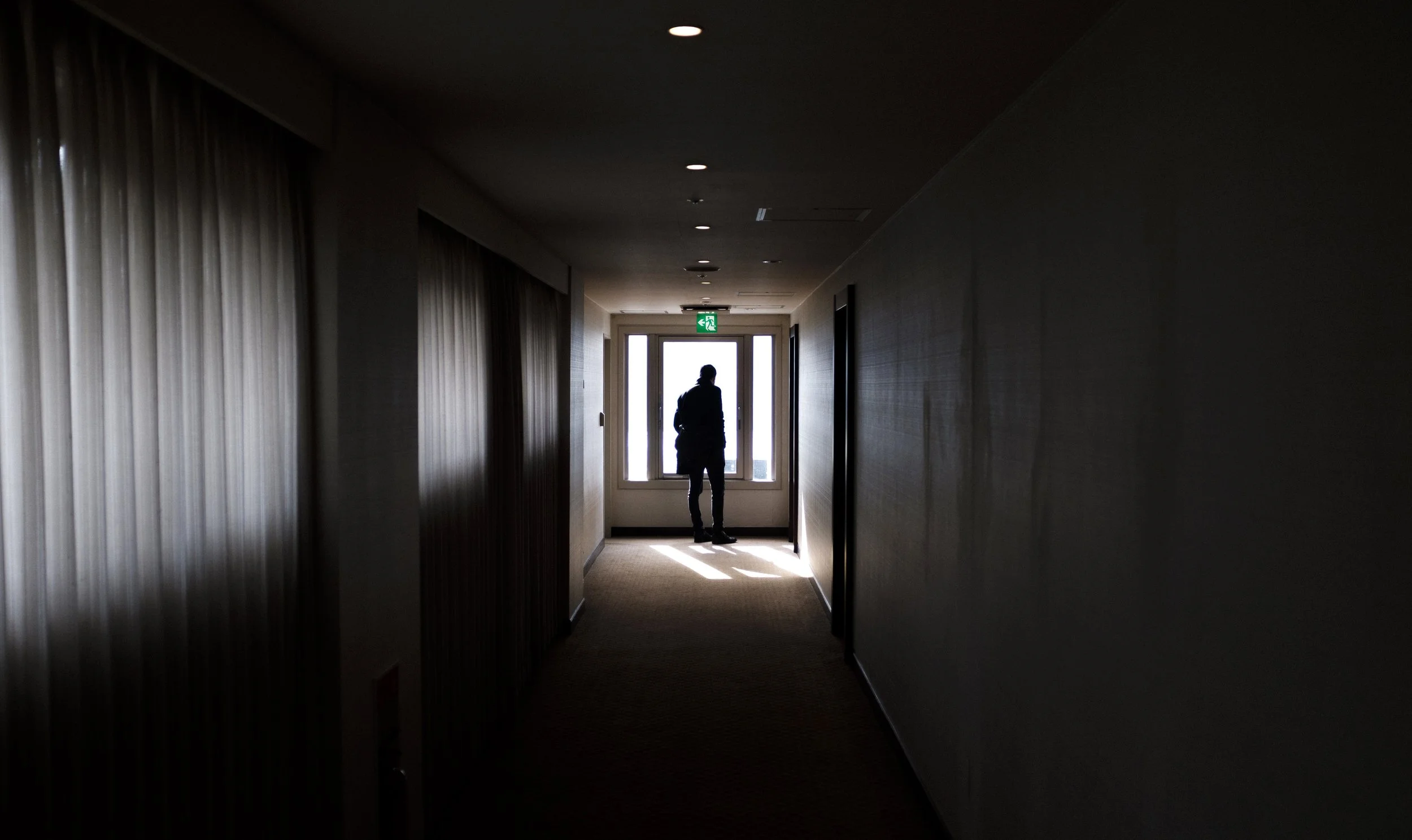Silhouette d'une personne debout près d'une porte-fenêtre à la fin d'un couloir sombre, lumière du dehors illuminant partiellement l'intérieur.