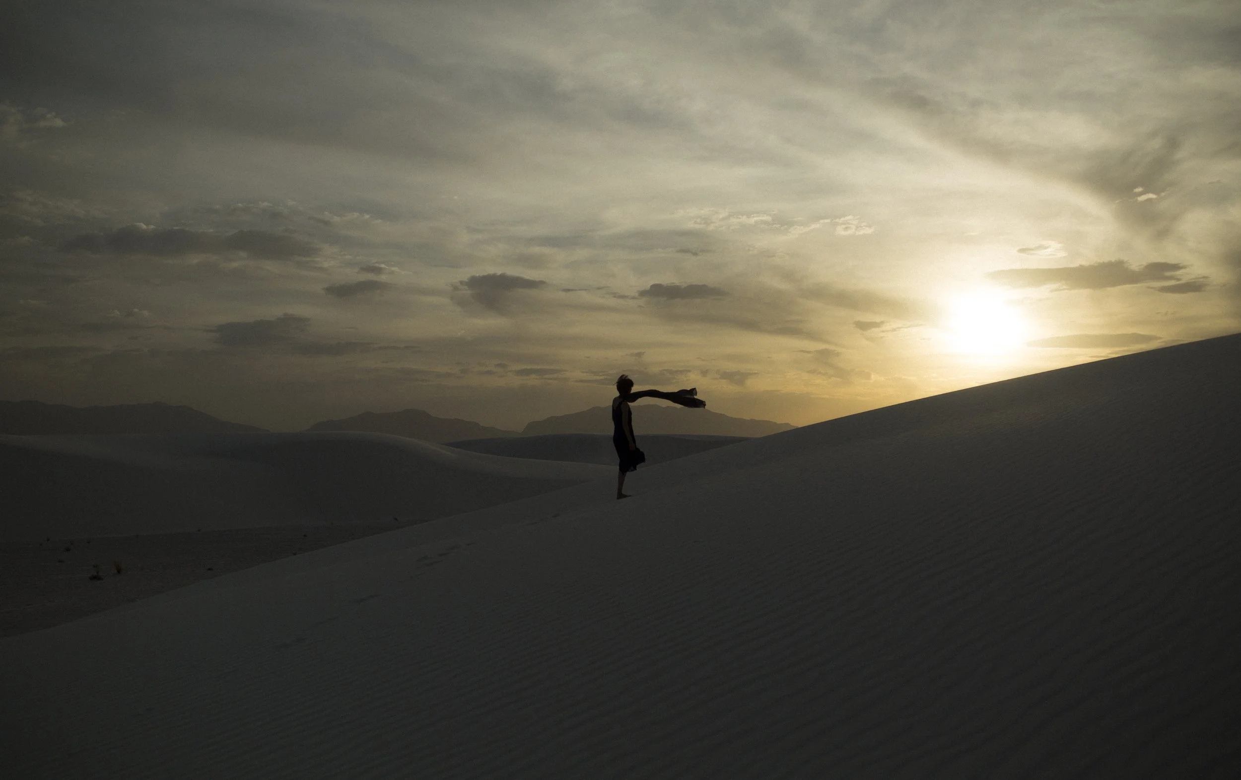Une personne se tient dans un désert de dunes, au coucher du soleil, avec un drapeau dans la main, créant une silhouette contre un ciel nuageux.