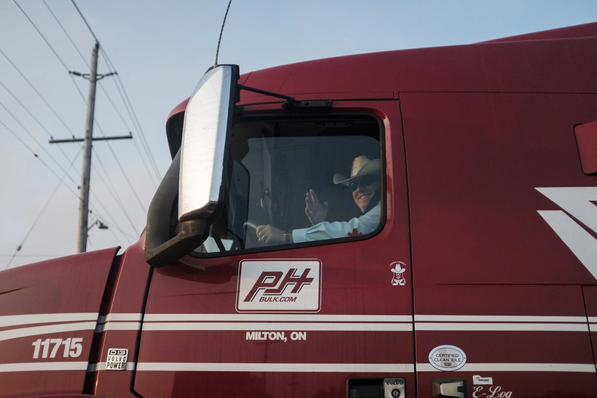 Un camion rouge avec un homme portant un chapeau cowboy et des lunettes, souriant et faisant un geste de la main, assis dans la cabine.