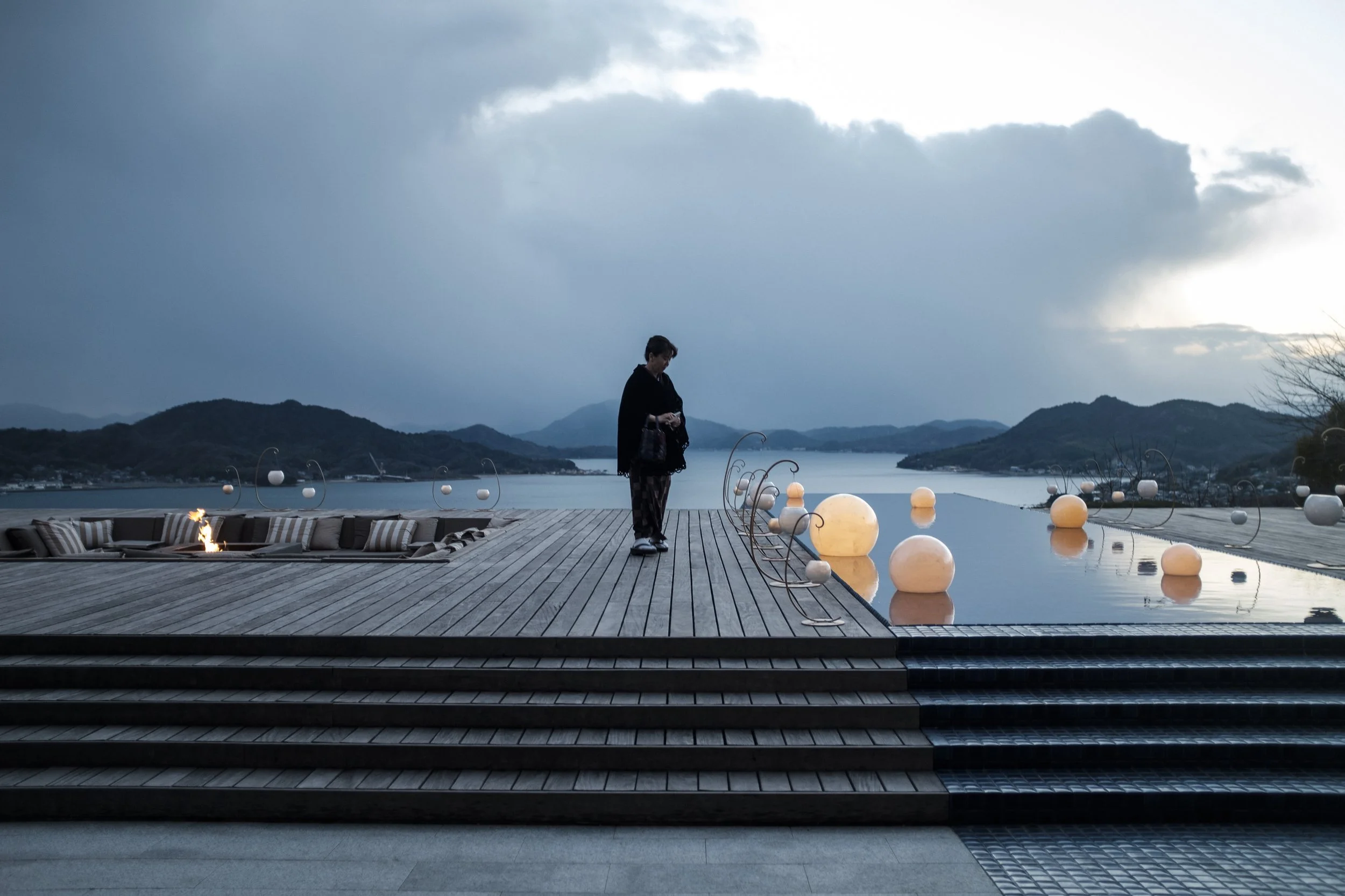 Une personne en vêtements noirs se tient près d'une piscine sur une terrasse en bois, avec des boules lumineuses flottant dans l'eau, un ciel nuageux et un paysage de montagnes en arrière-plan.