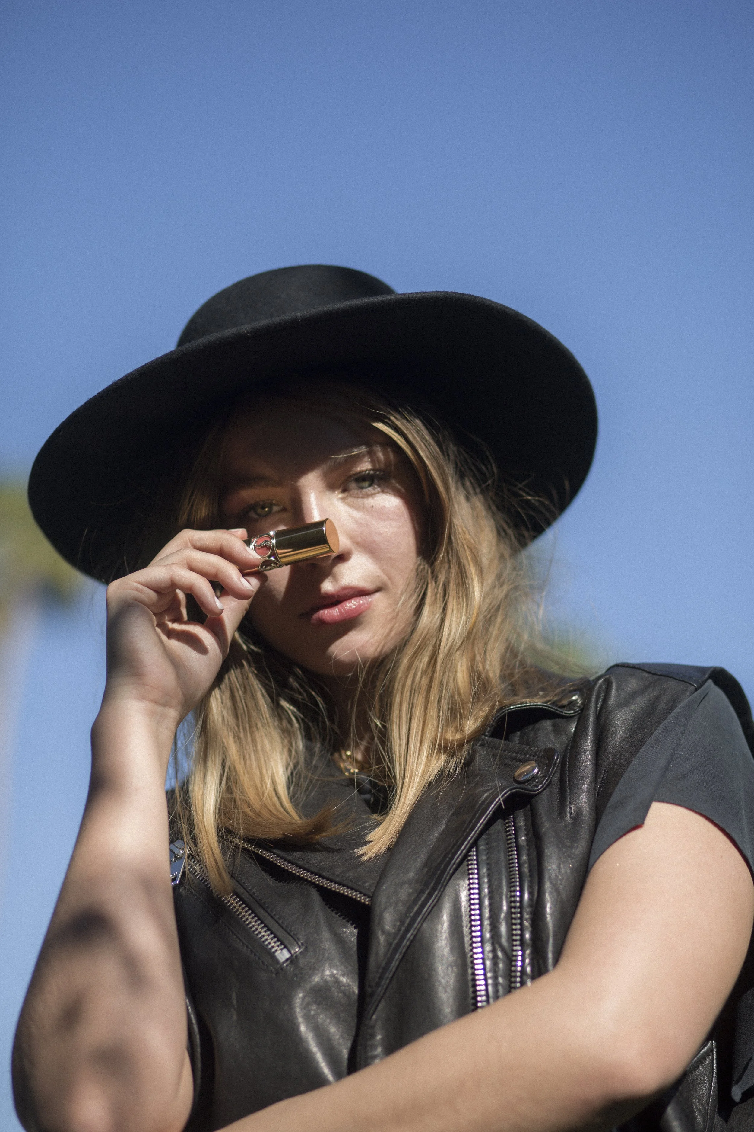 Une femme portant un chapeau noir et une veste en cuir, tenant un petit objet doré près du nez, avec un ciel bleu en arrière-plan.