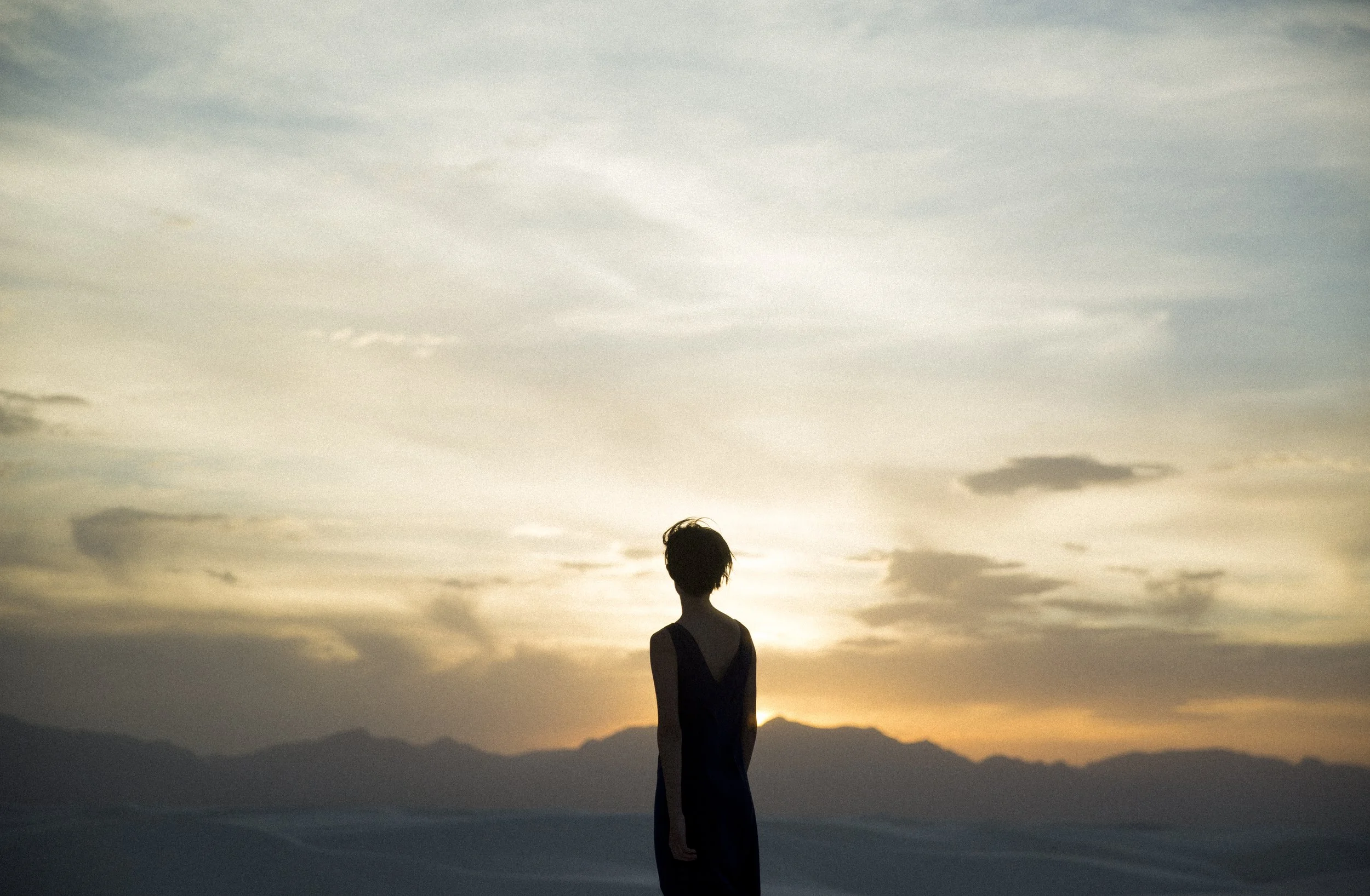 Une silhouette de femme en robe noire debout devant un coucher de soleil, avec des montagnes en arrière-plan.