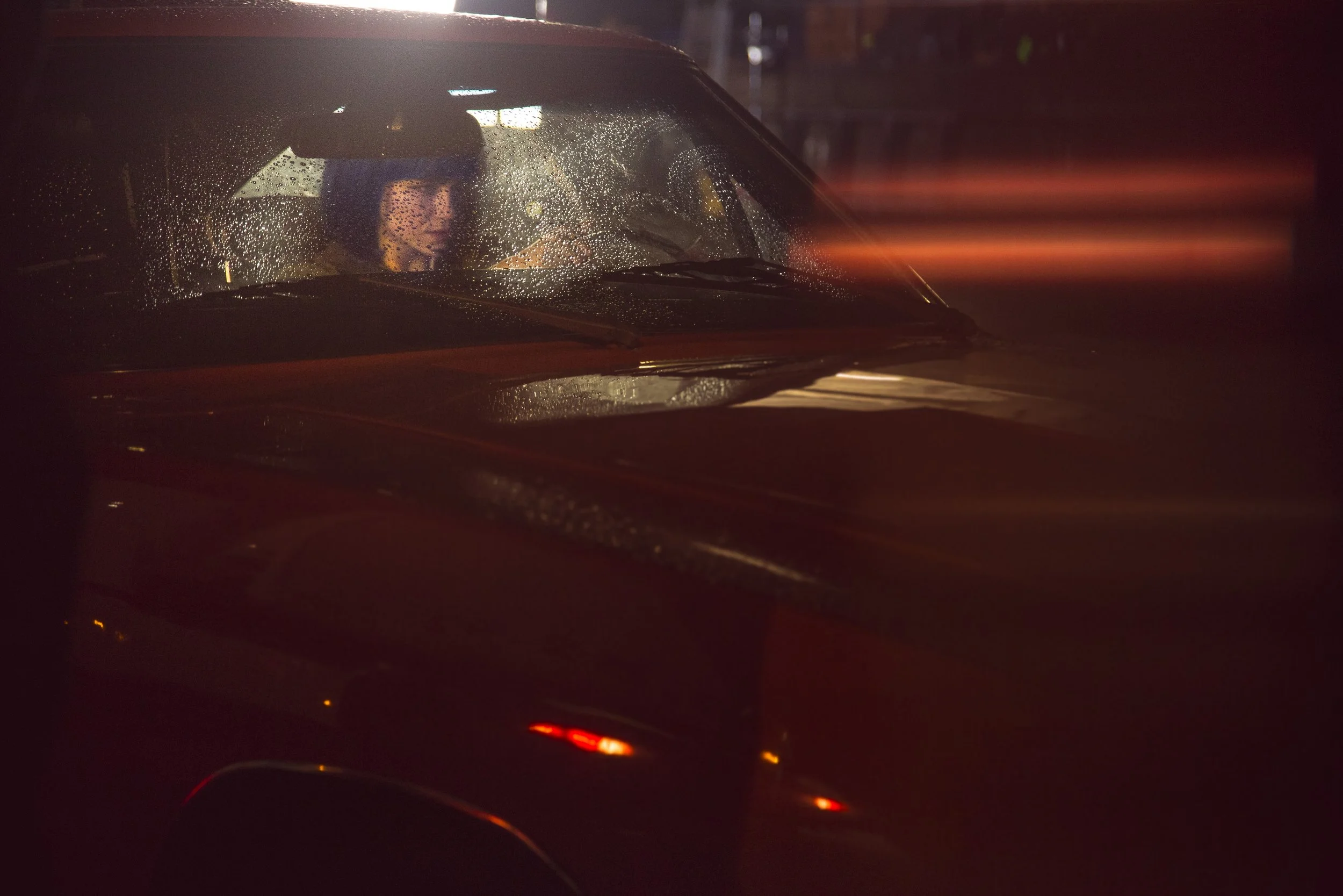 Voiture noire vue de face la nuit, avec des gouttes de pluie sur le pare-brise et des reflets rouges d’éclairages.