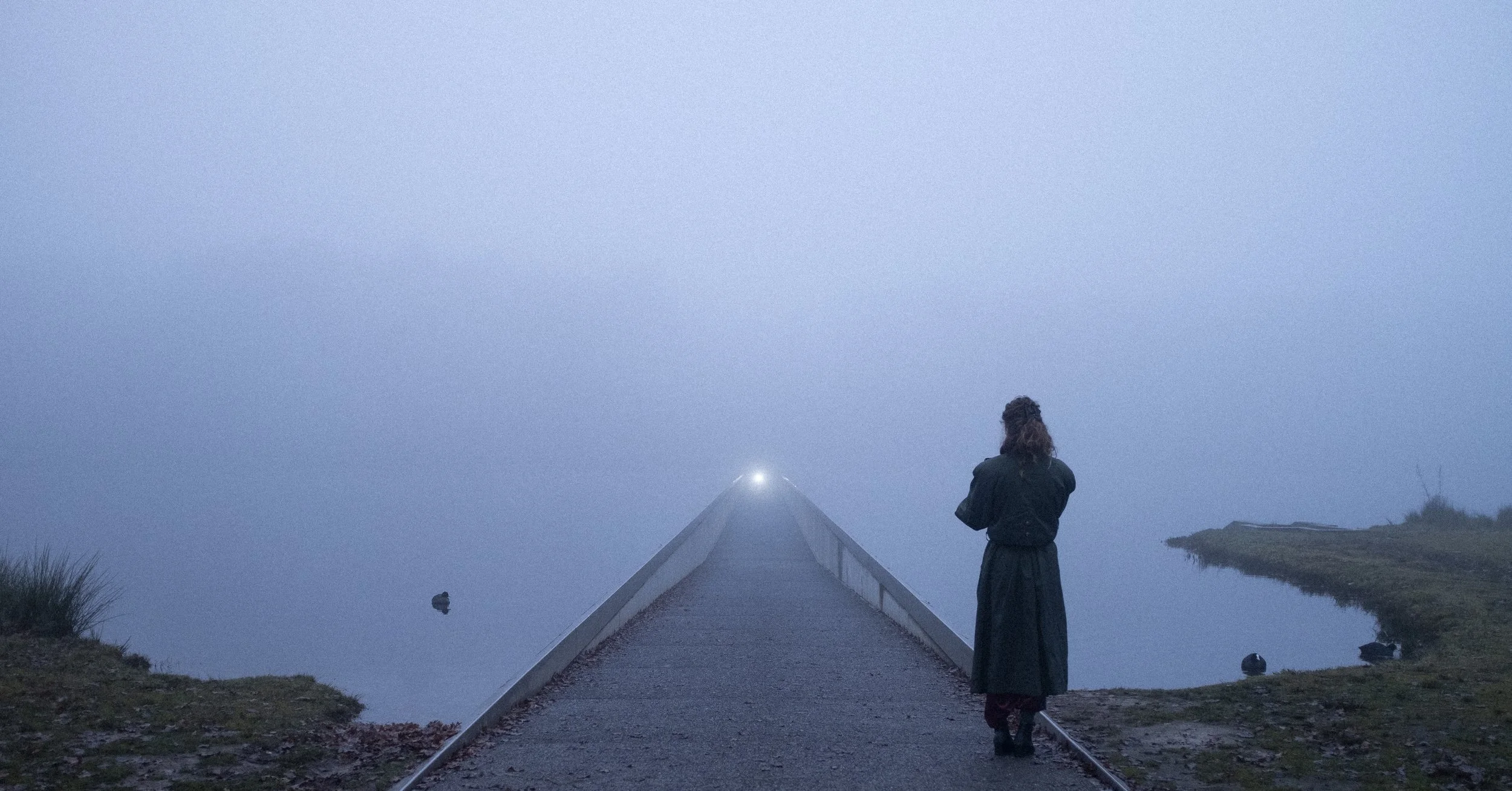 Une personne habillée en vêtement sombre regarde au loin sur un pont en pleine brume, avec une étendue d'eau de chaque côté.