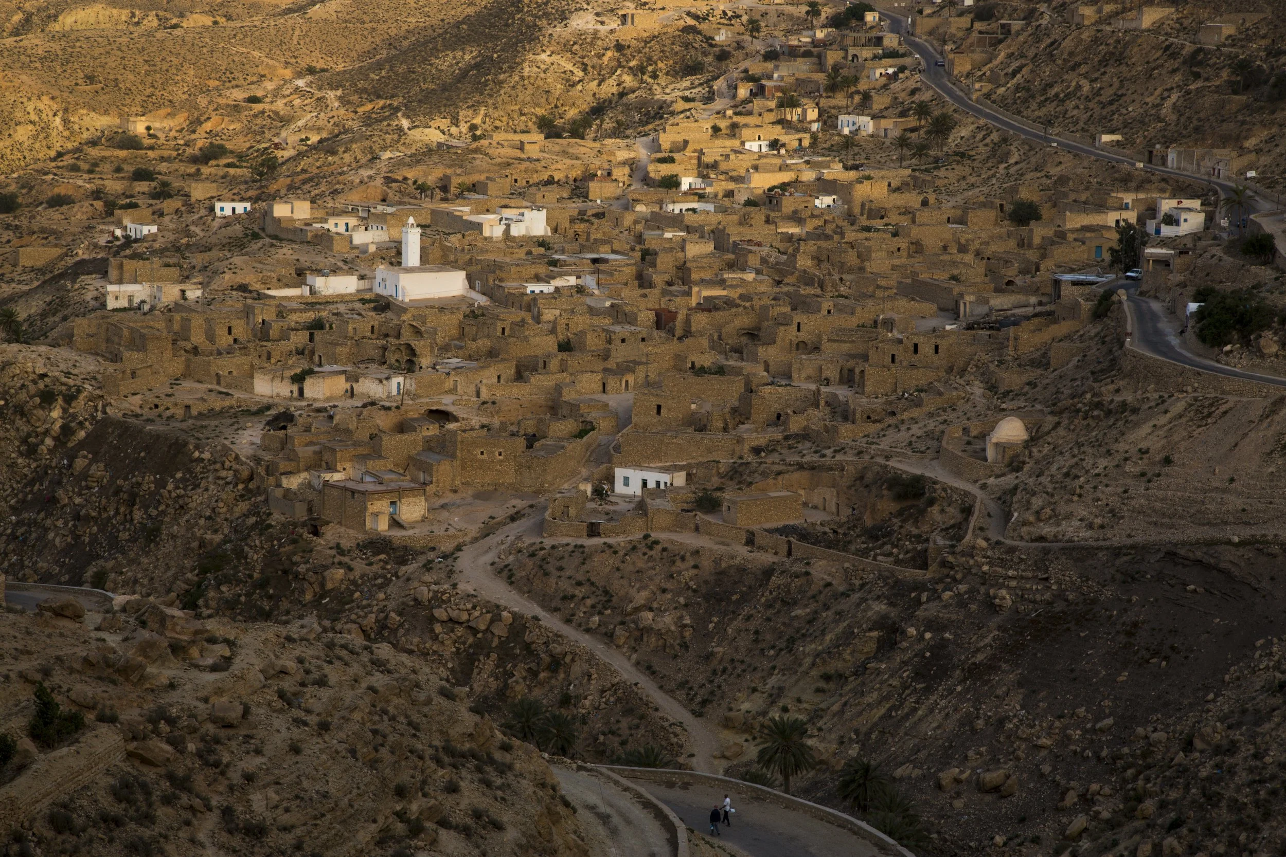 Un village berbère construit sur une colline rocheuse, avec des maisons en pierre beige et plusieurs bâtiments blancs, entouré d'un paysage désertique montagneux, avec une route sinueuse traversant la vallée et deux personnes marchant.