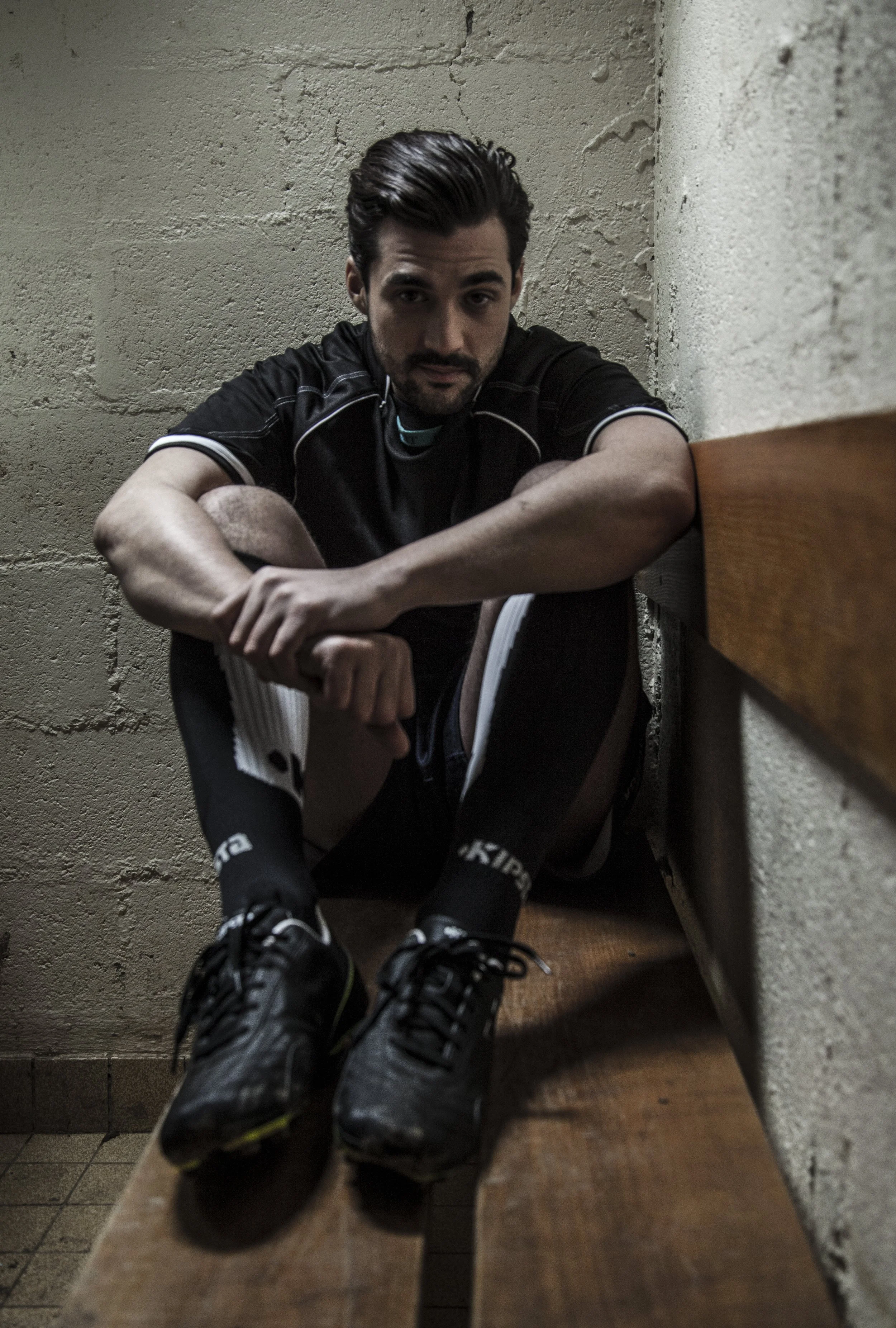 Un homme en vêtements de sport assis sur un banc dans une pièce aux murs en béton, regardant vers l'objectif.