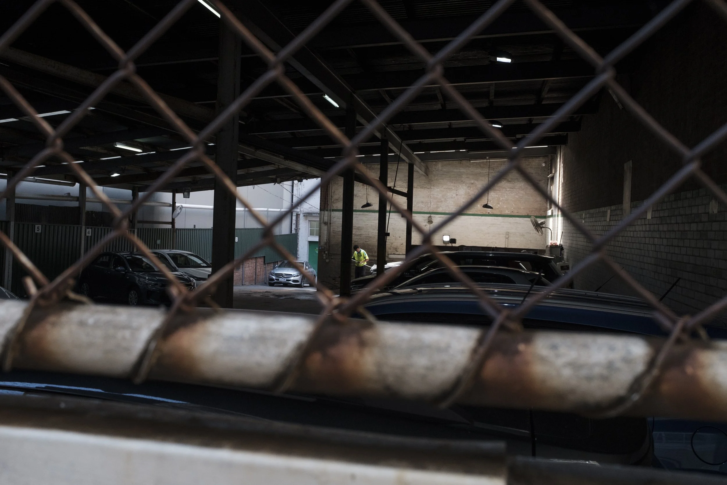 Vue d'un parking souterrain avec des voitures, vue à travers une grille rouillée, un homme en veston jaune dans le fond qui travaille.