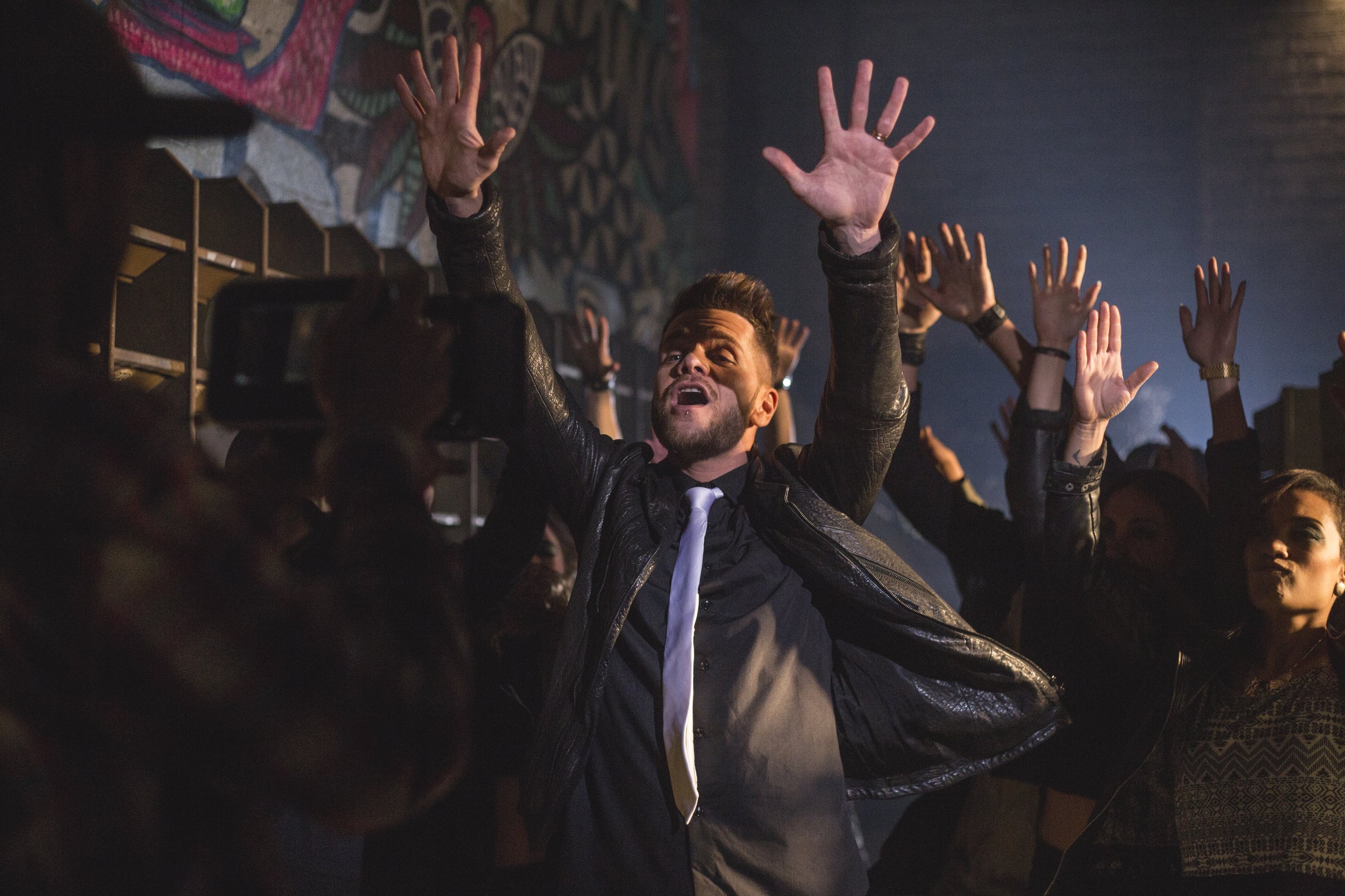 Un groupe de personnes en train de danser lors d'une soirée dans une ambiance sombre avec des éclairages tamisés, au centre un homme avec une barbe portant une chemise noire, une cravate blanche et une veste en cuir, a les bras levés et semble s'amus