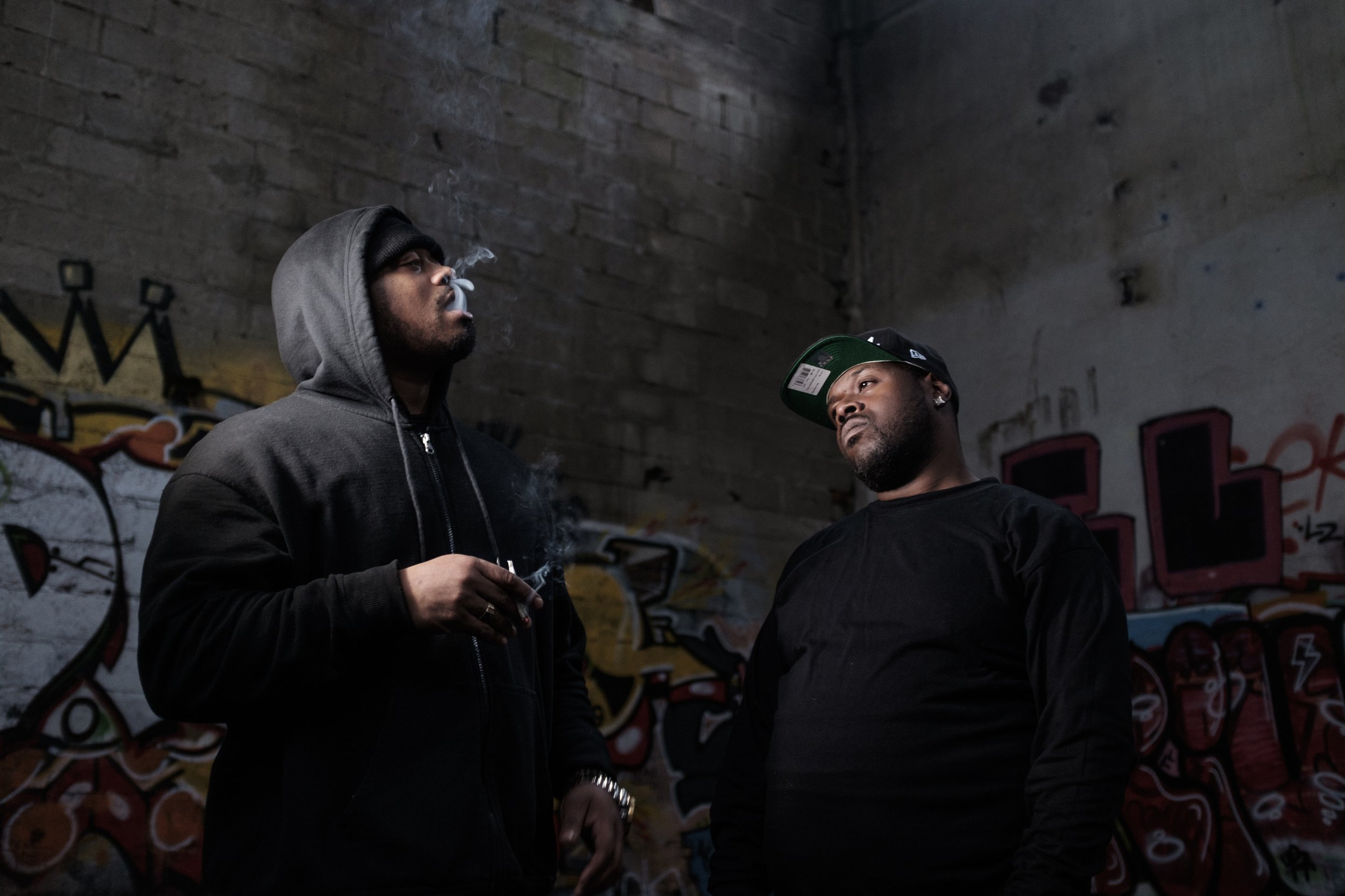 Deux hommes dans un sous-terrain graffité, l'un porte une capuche et fume, l'autre porte une casquette et regarde l'autre.