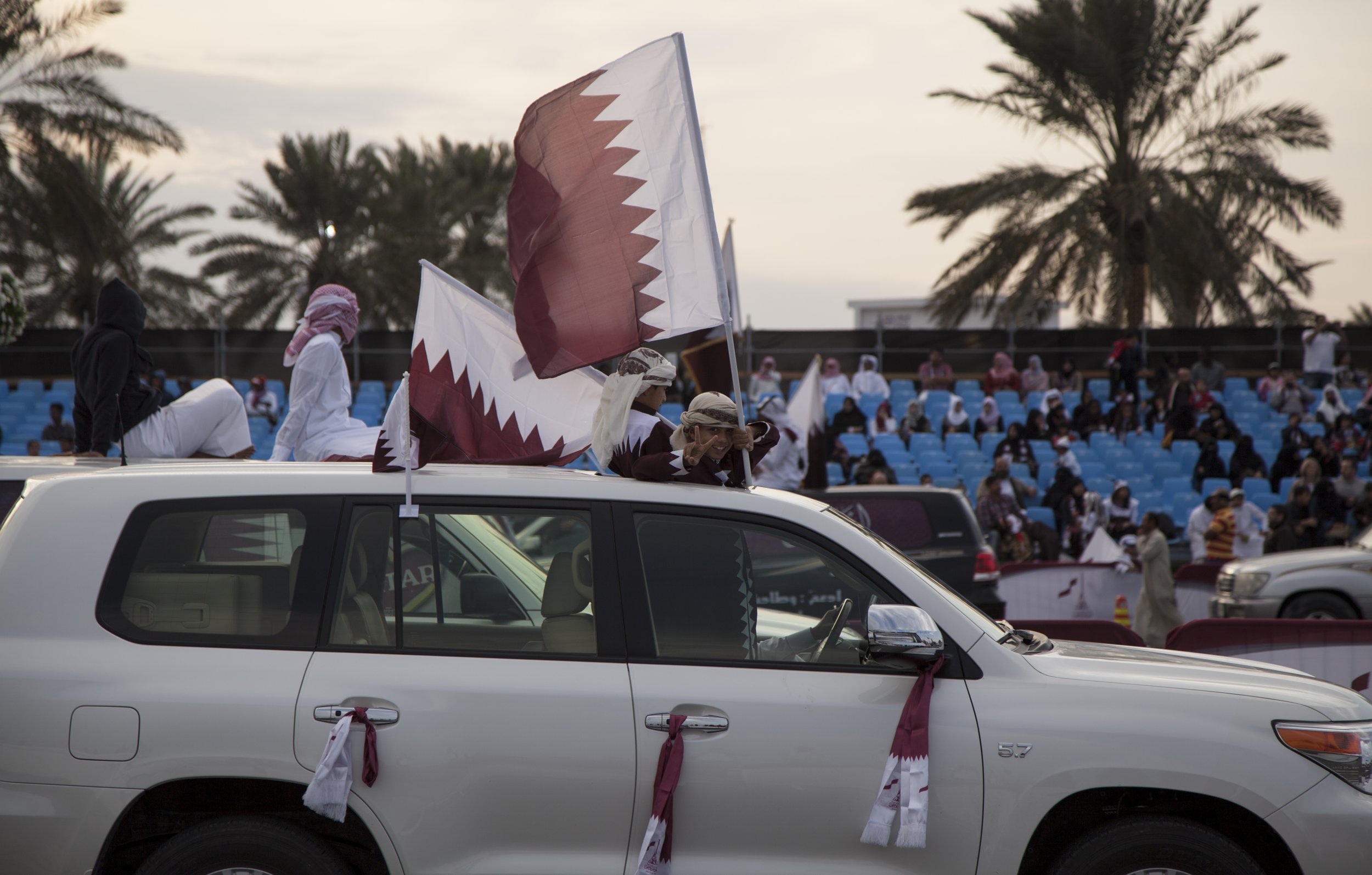Des personnes dans un véhicule blanc, décoré avec des foulards ou des tissus aux couleurs du Qatar, participent à une procession ou un événement, avec un public installé dans des gradins en arrière-plan.