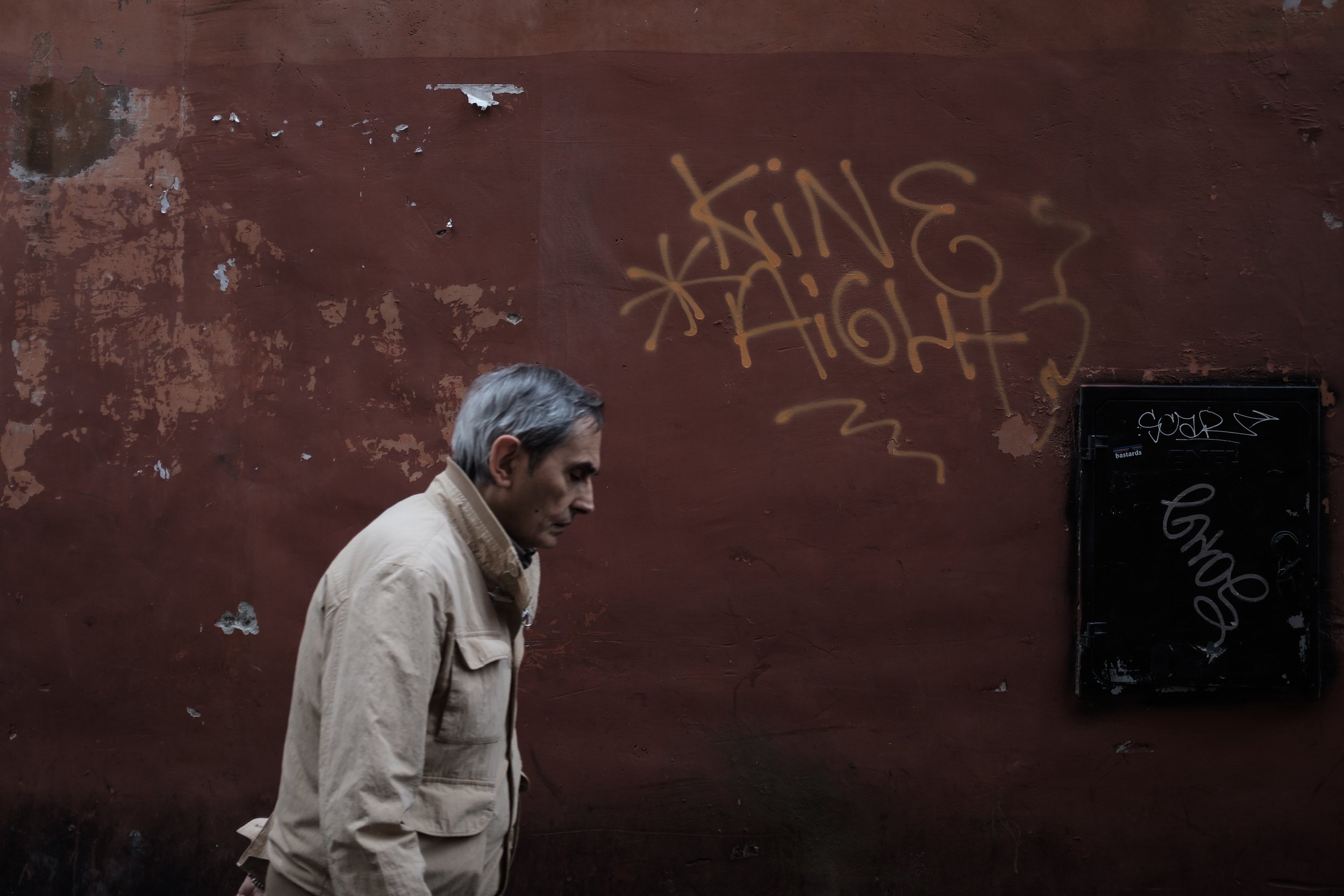 Un homme âgé avec des cheveux gris marchant devant un mur rouge avec des graffitis, dont l'un dit 'KING OF HIGH'.