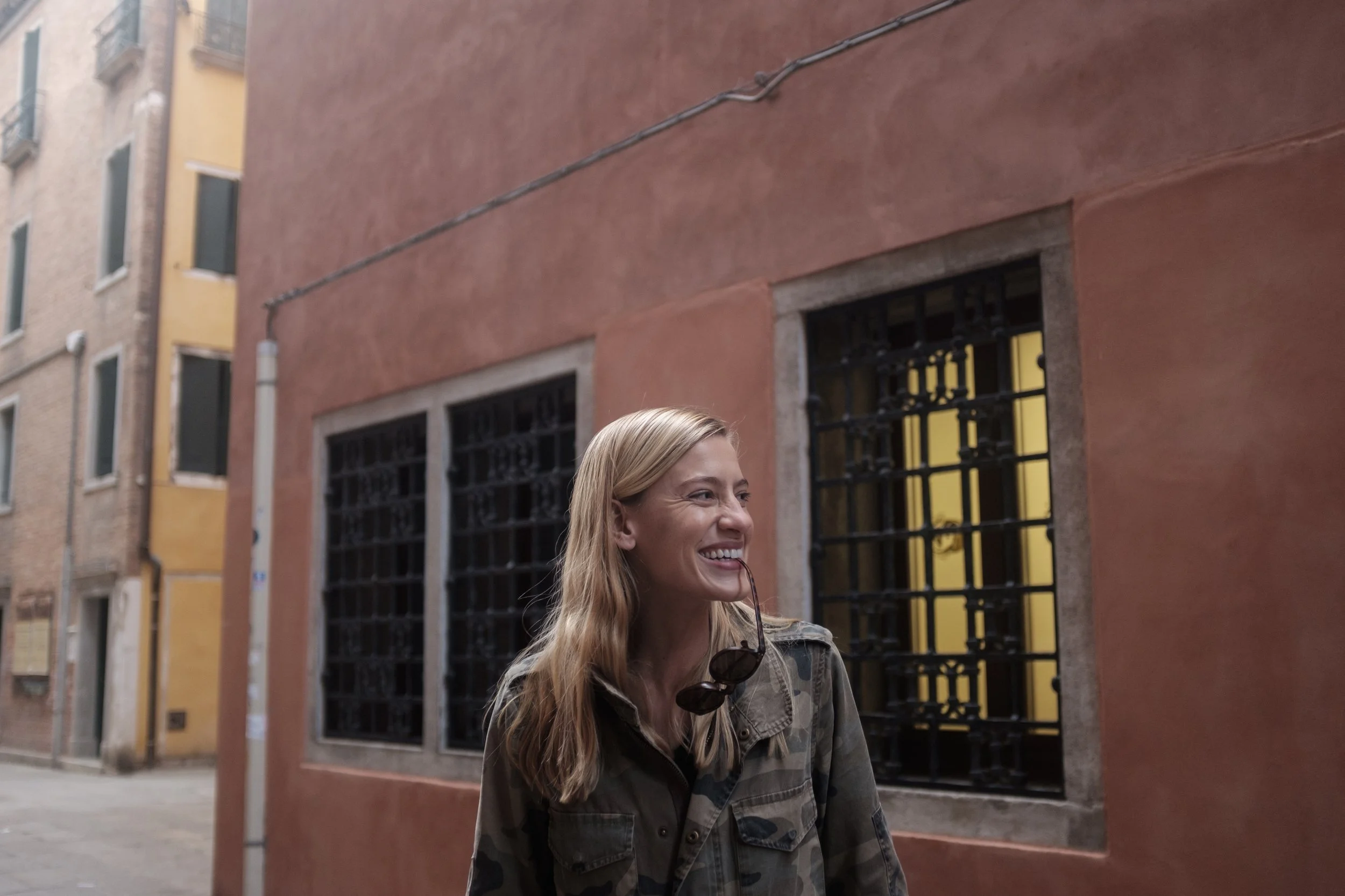 Femme souriante portant une veste camouflage, tenant des lunettes de soleil, devant un bâtiment en briques avec fenêtres grillagées.