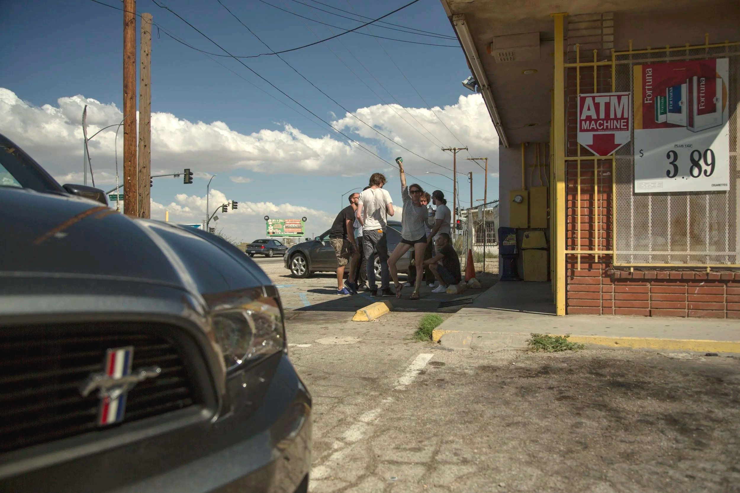 Groupe de jeunes femmes et hommes en train de discuter à la sortie d'un magasin à proximité d'une station-service, avec des voitures garées dans le parking et un ciel orageux en arrière-plan.
