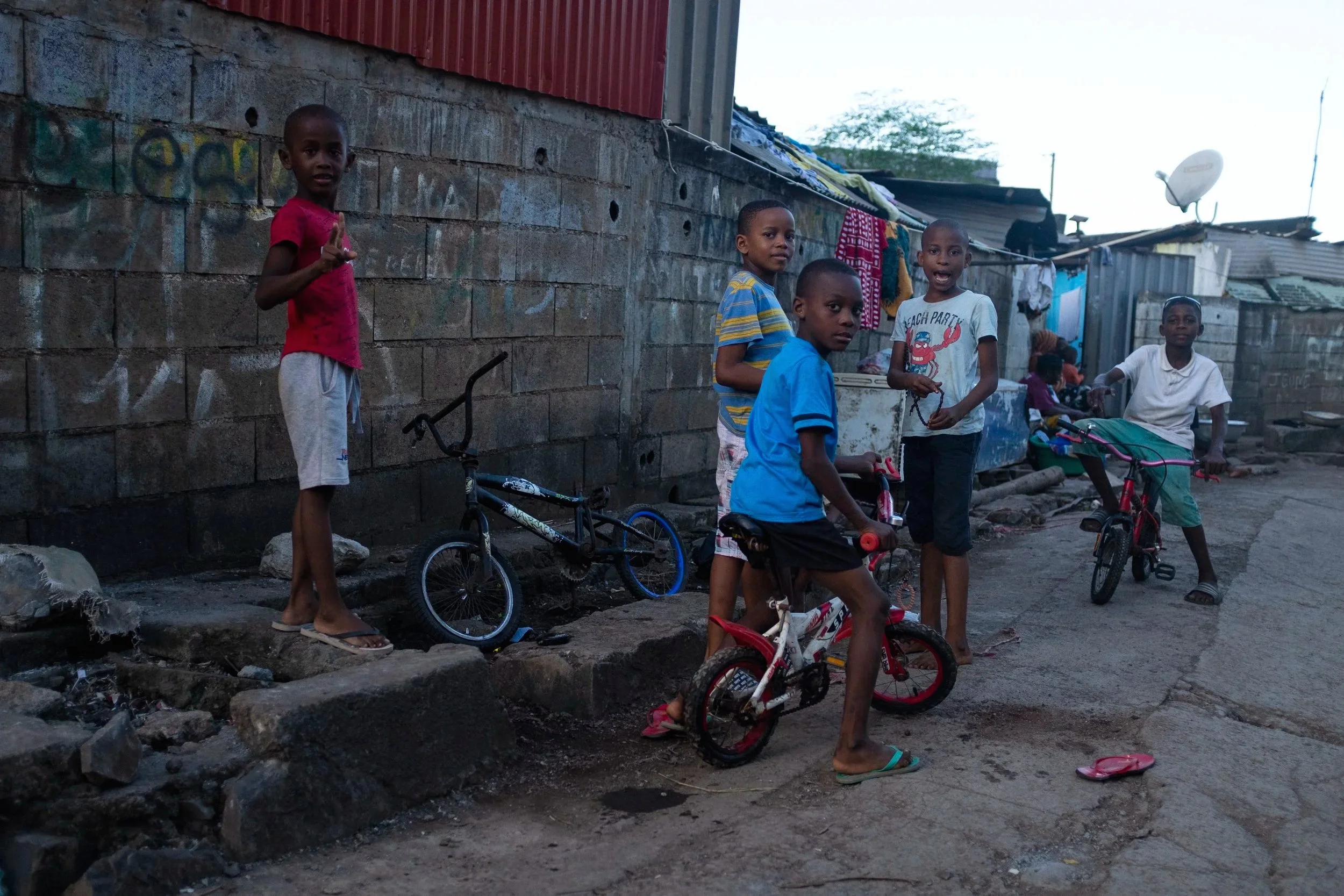 Enfants jouant avec des vélos dans une rue d'une zone urbaine modeste, avec des murs en briques et des habitations en tôles ondulées en arrière-plan.