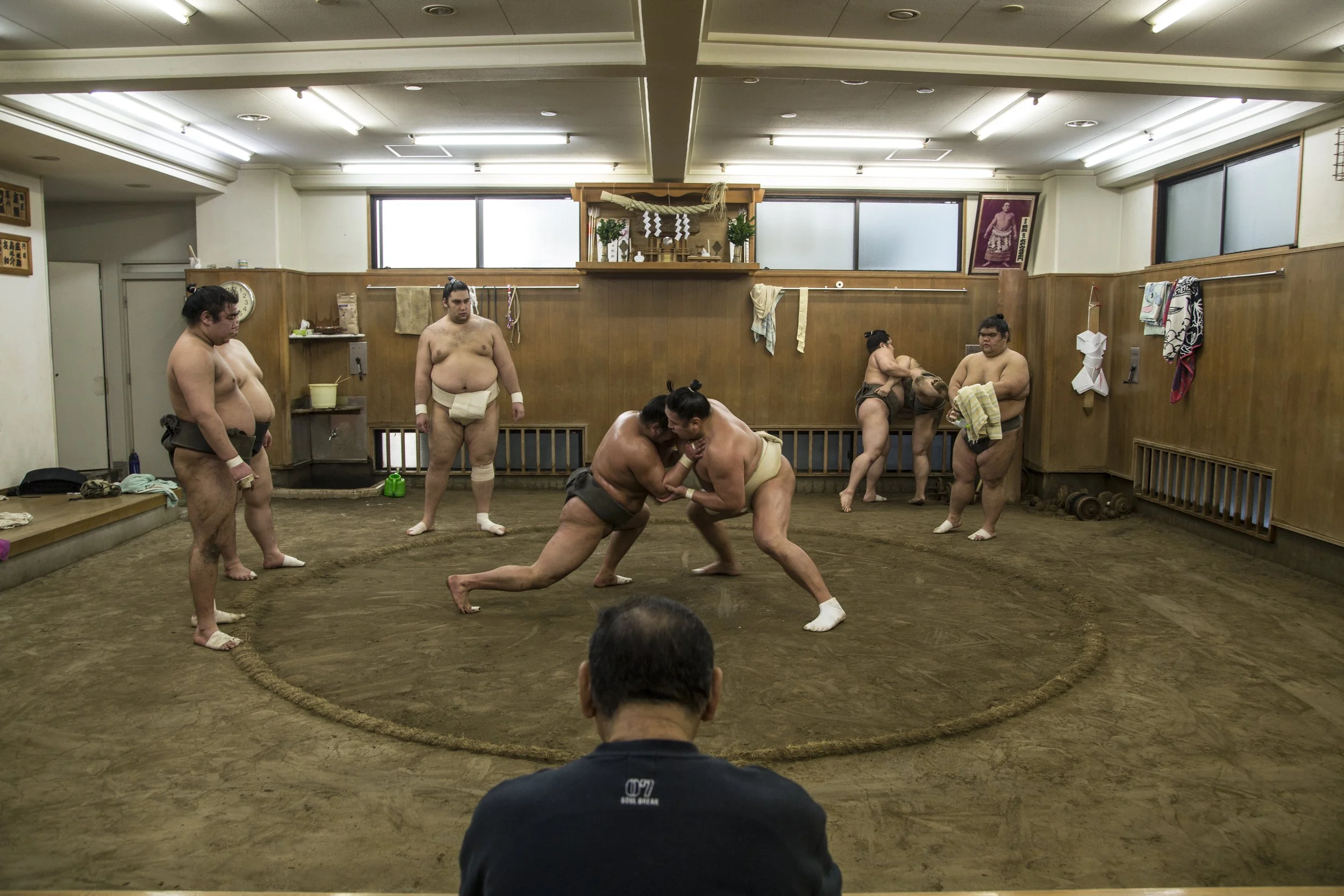 Une scène de sumo dans une salle en bois avec deux lutteurs en pleine confrontation, plusieurs autres lutteurs en attente et un spectateur en arrière-plan.