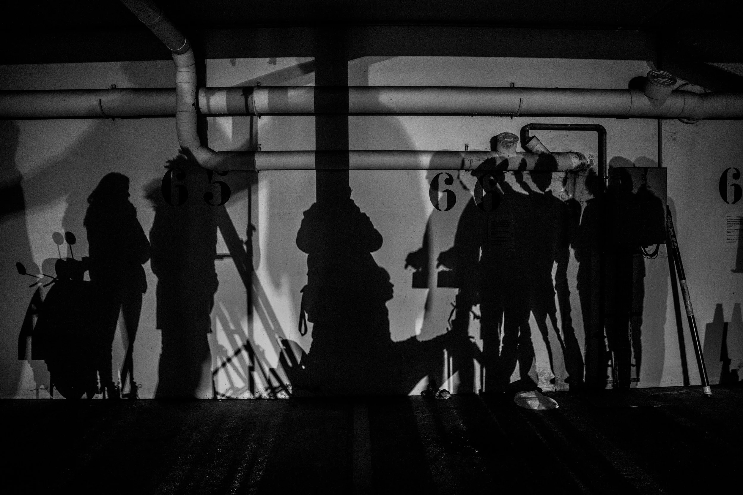 Ombres de plusieurs personnes avec des vélos et une pelle, projetées contre un mur dans un parking souterrain, en noir et blanc.