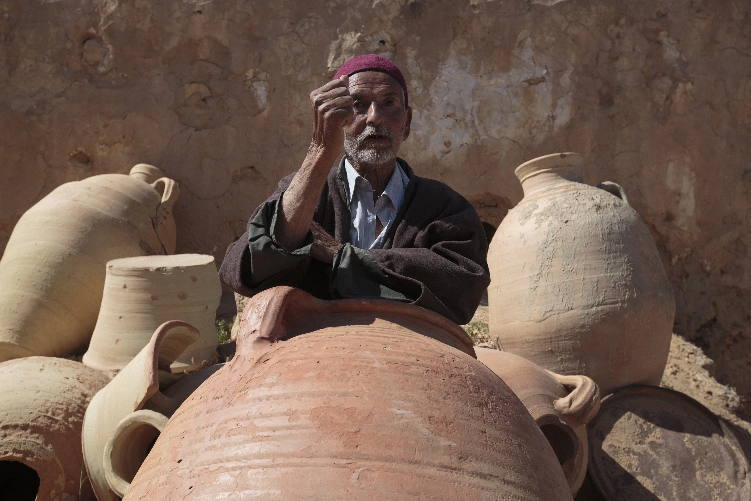 Un homme âgé, portant une casquette rouge, est assis parmi de grands pots en terre cuite, dans un environnement en pierre.