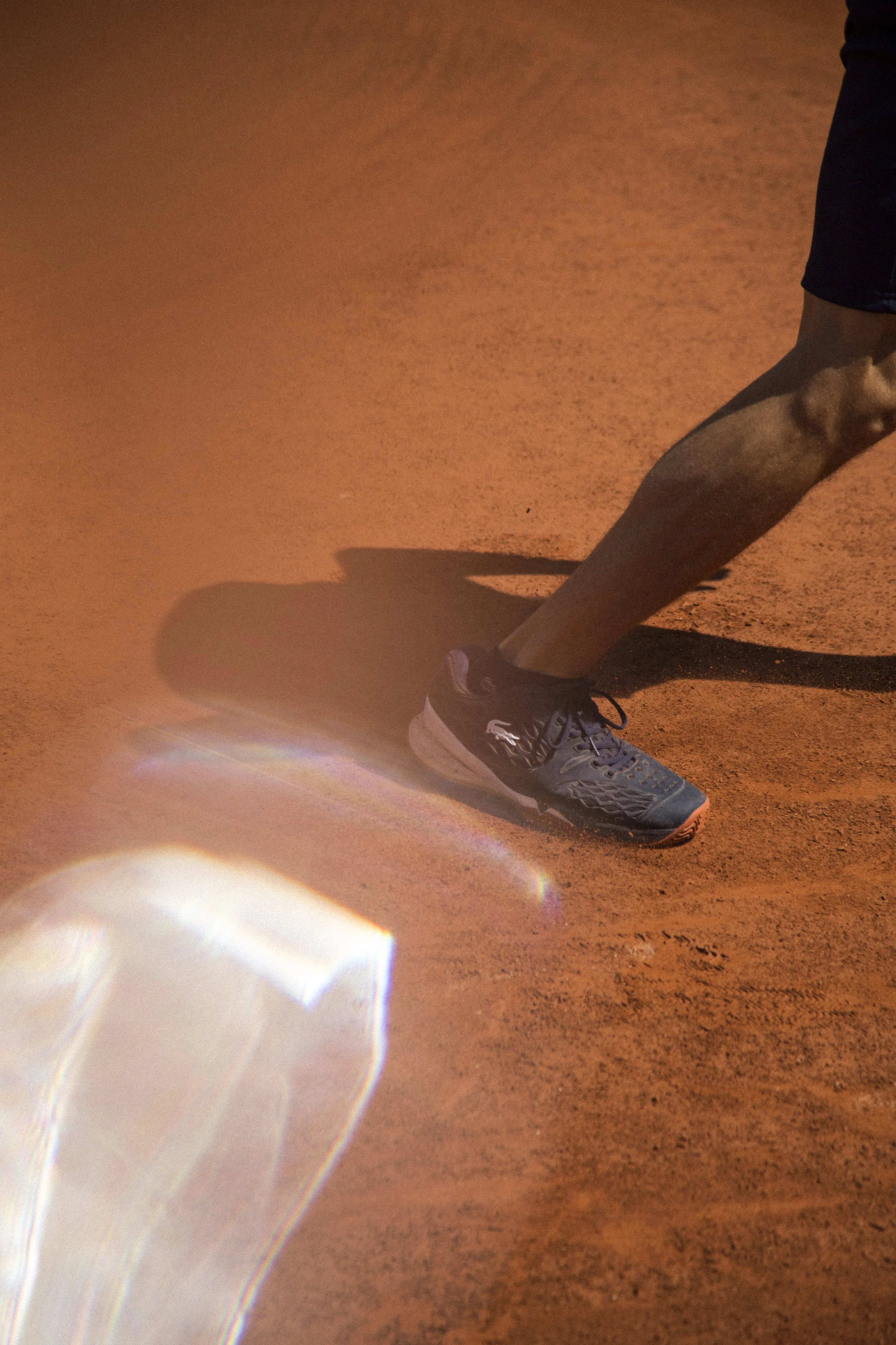 L'image montre une personne jouant au tennis sur un court en terre battue, portant des chaussures de sport et un short, avec une ombre prononcée sur le sol.
