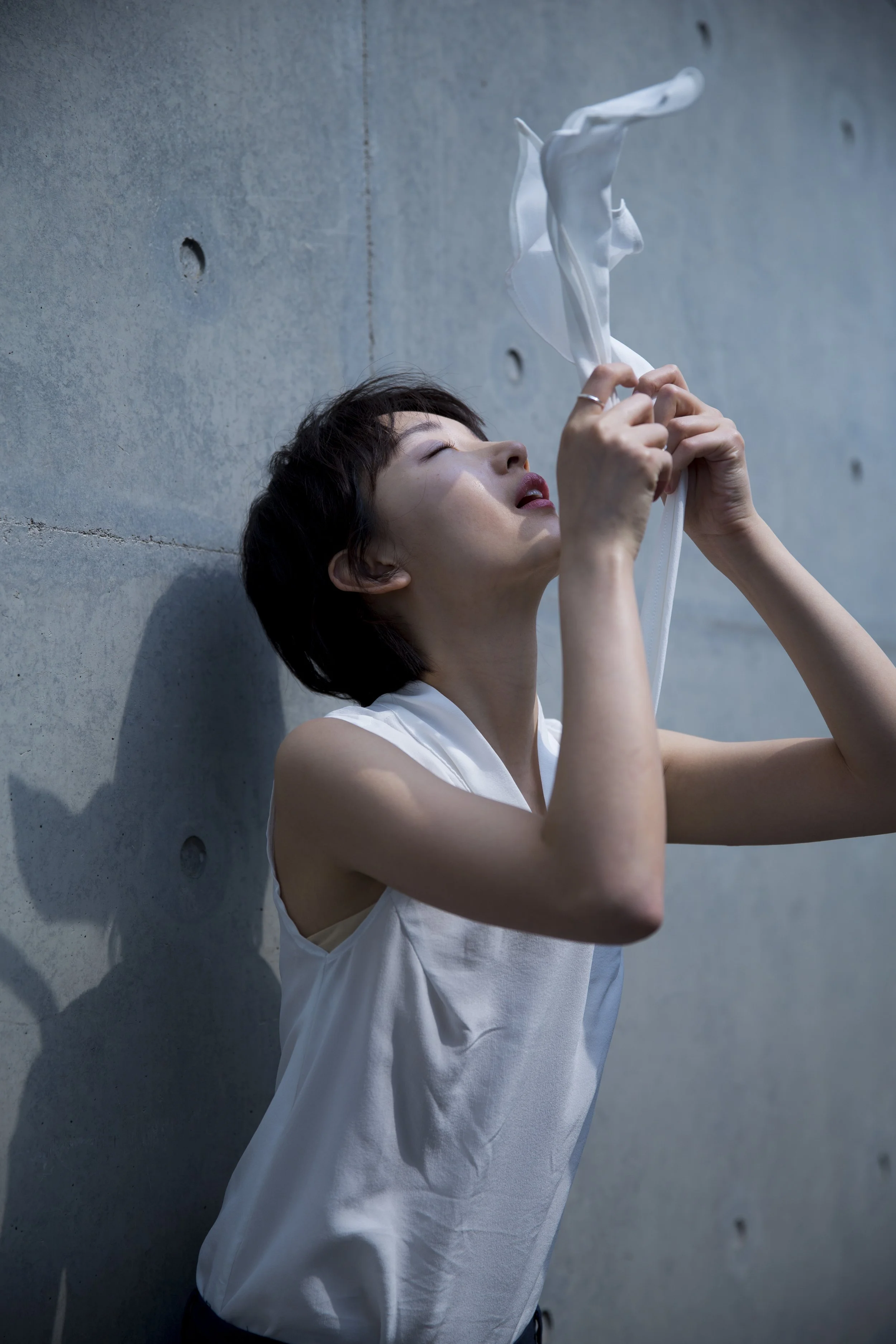 Une femme avec des cheveux court, portant une chemise blanche, appuyée contre un mur en béton, tenant un morceau de tissu blanc