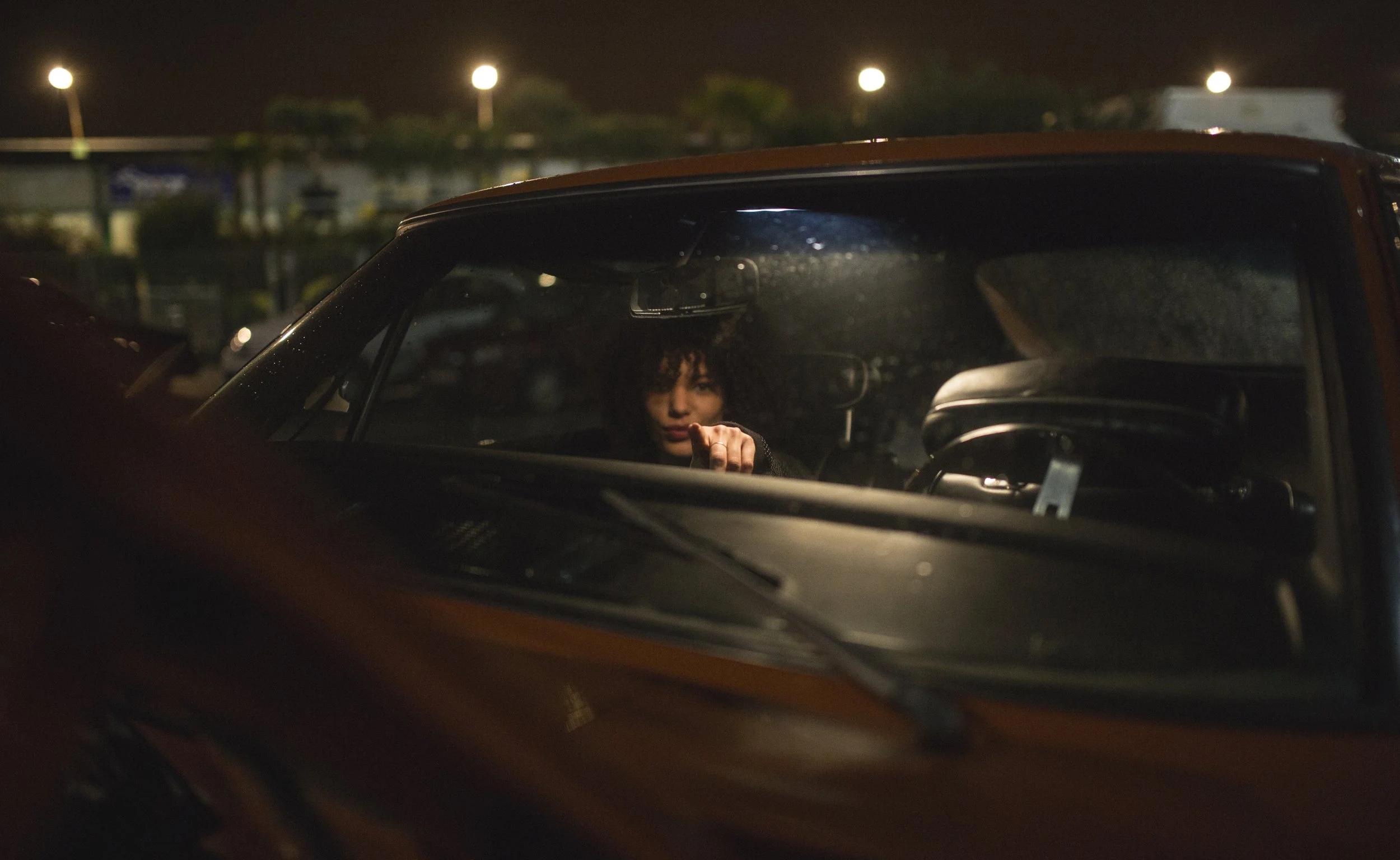 Une femme avec des cheveux frisés pointant du doigt à travers le pare-brise d'une voiture sombre, la scène se déroule la nuit dans un parking.