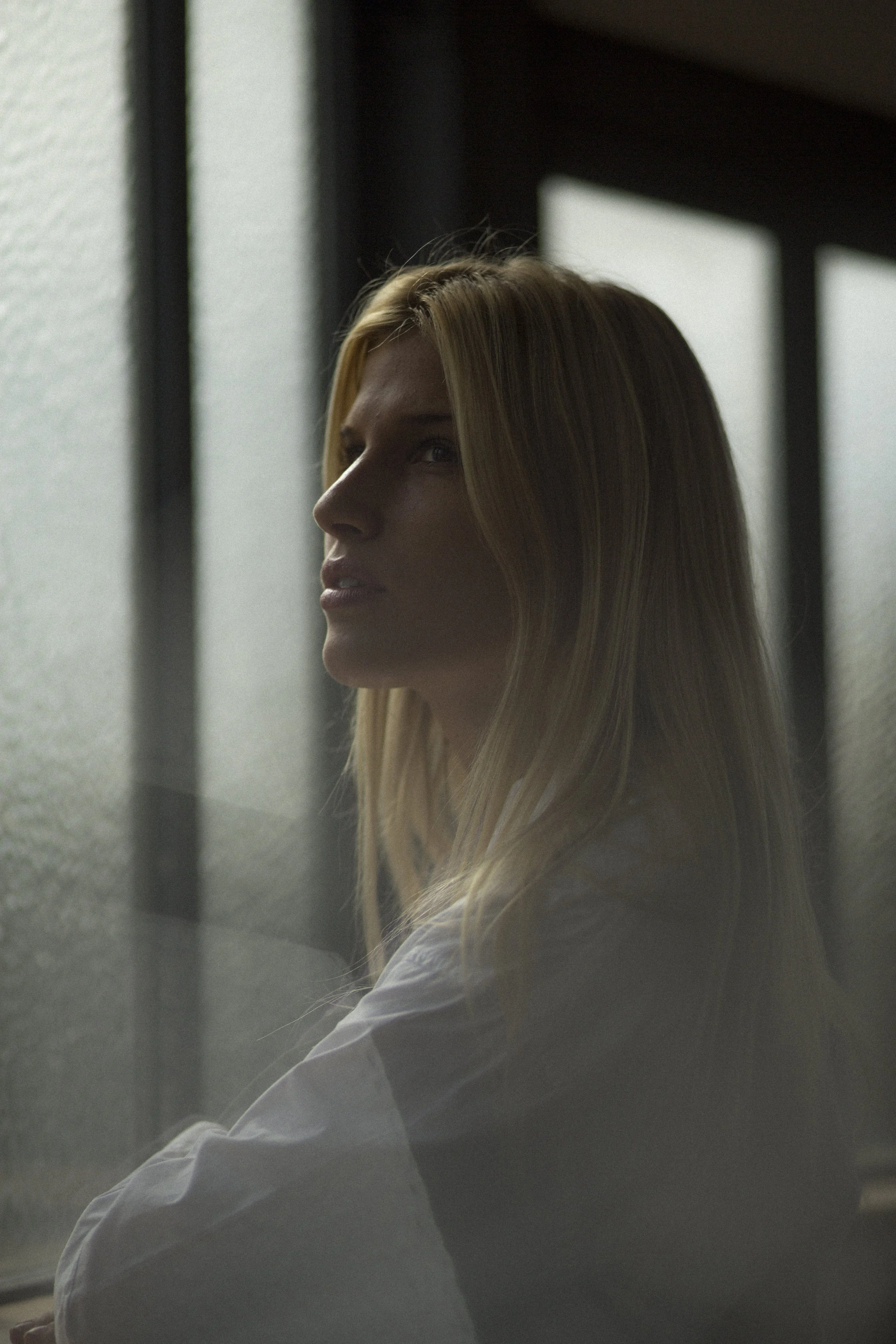 Une femme aux cheveux blonds regarde par une fenêtre avec une expression pensive, dans une ambiance sombre et calme.