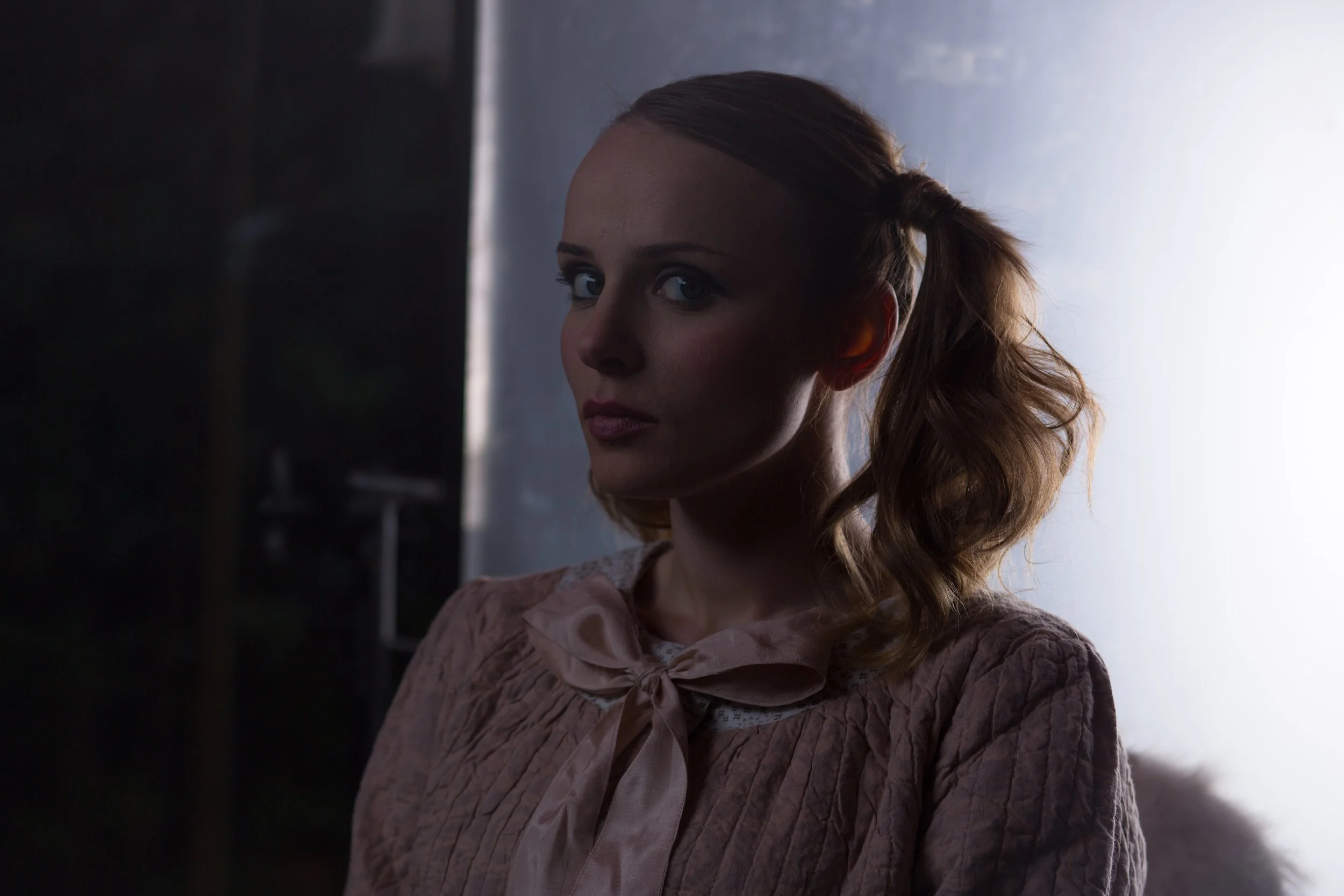Une femme aux cheveux bouclés en queue de cheval regarde vers la caméra dans un éclairage tamisé.