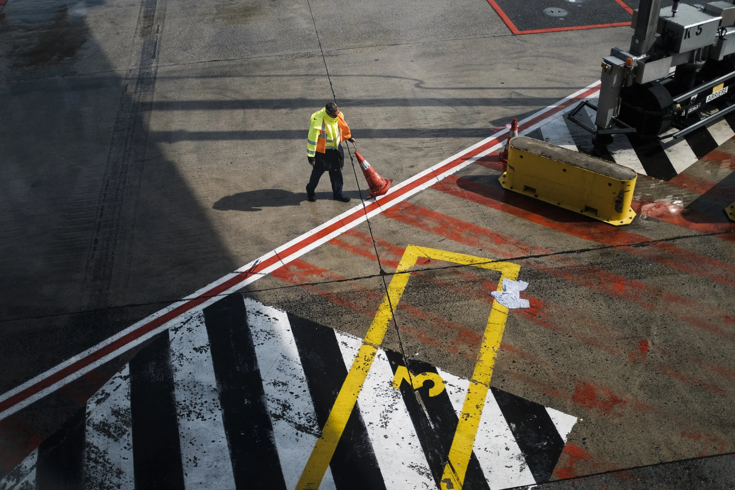 Un travailleur en gilet réfléchissant jaune et orange, posant un cône orange sur une zone de piste d'aéroport, avec des marquages au sol en blanc, noir et jaune, et un équipement de sécurité en arrière-plan.