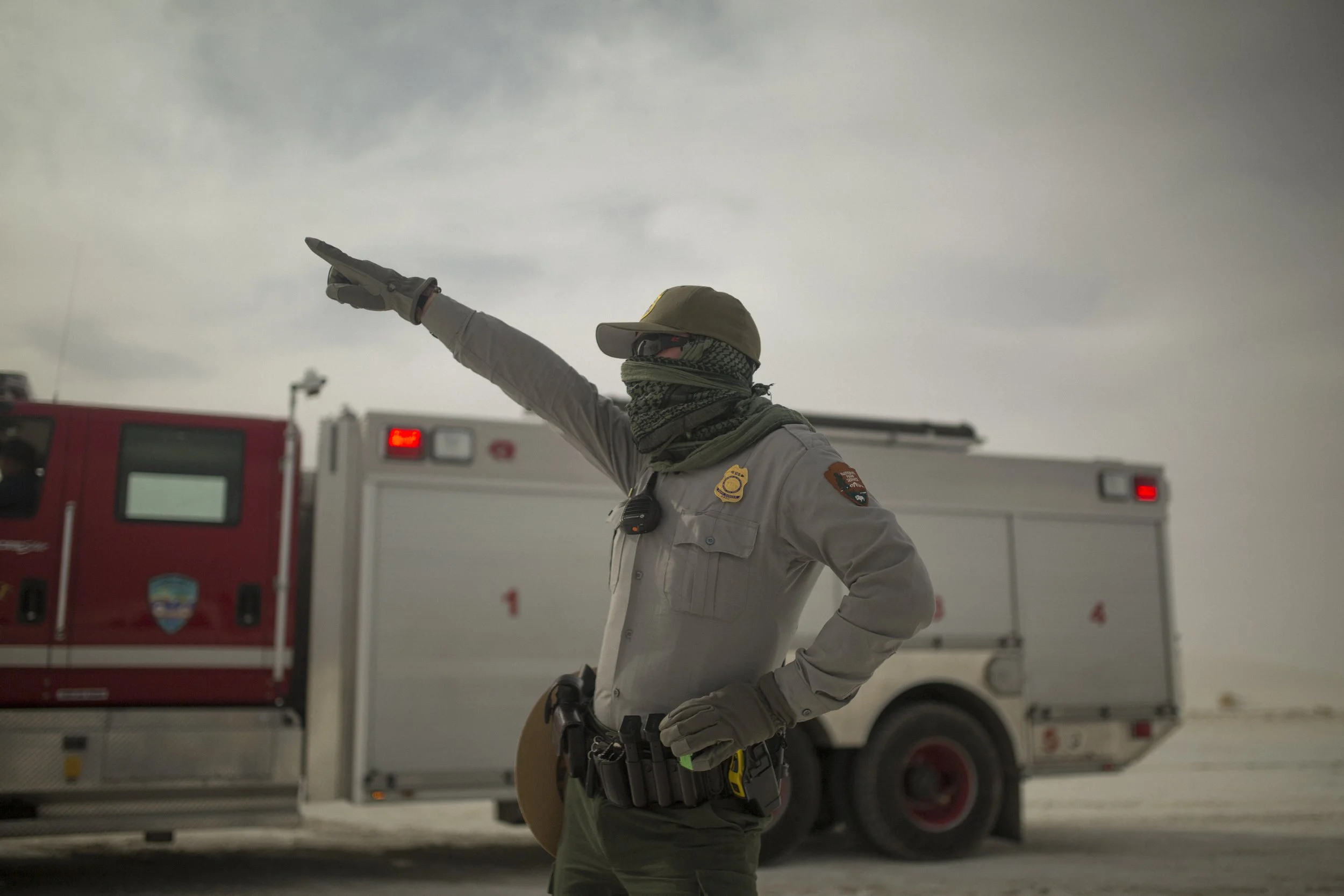Un agent de sécurité ou un détective avec un casque, lunettes de soleil, gilet pare-balles, et gants, pointant du doigt devant un véhicule de secours ou de lutte contre les incendies dans un environnement désertique ou en zone aride.