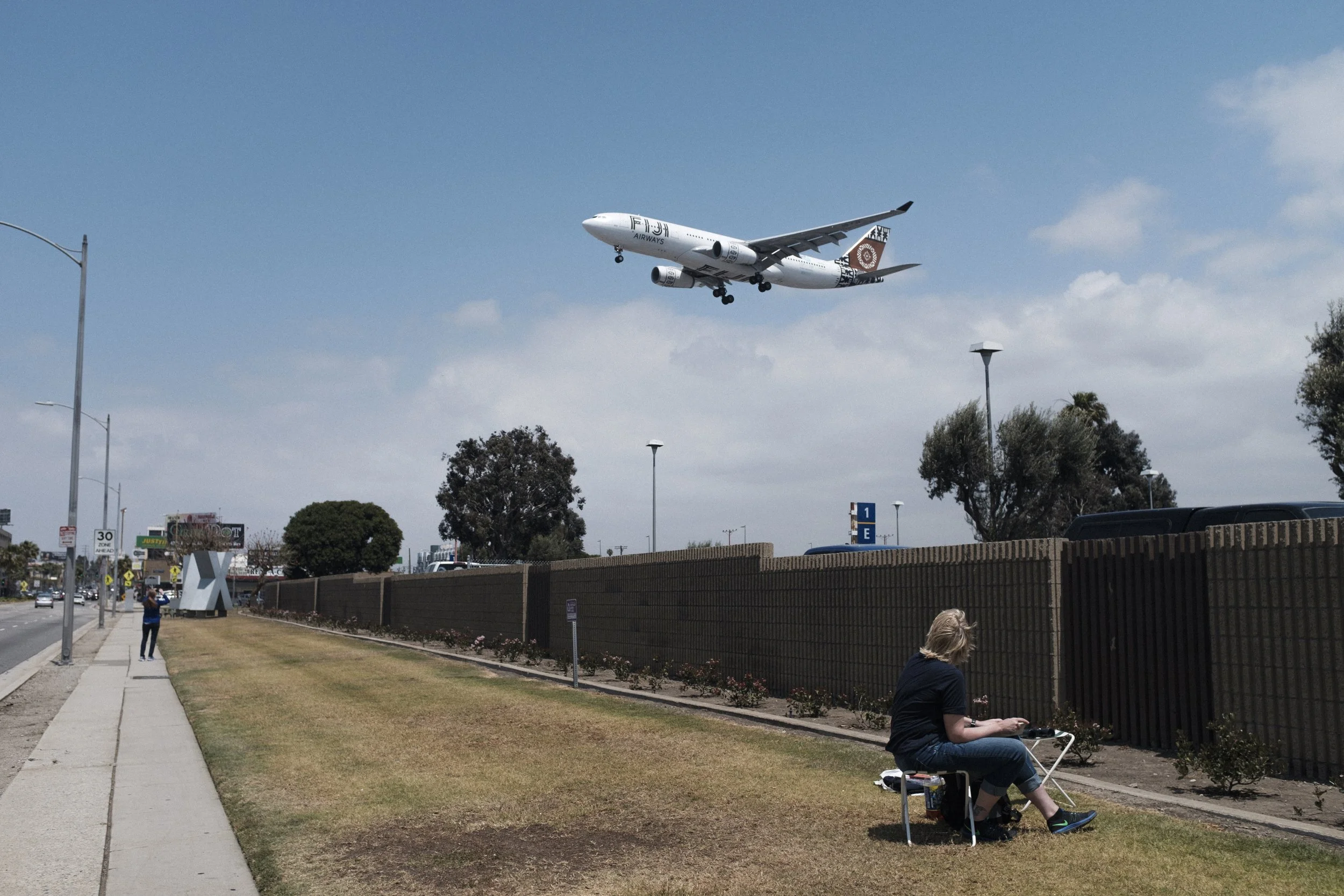 Un avion en approche d'atterrissage dans le ciel bleu au-dessus de la ville, avec quelques nuages, alors qu'une femme est assise sur une chaise pliable près d'une clôture en attendant l'avion, et une personne marche sur le trottoir à gauche.